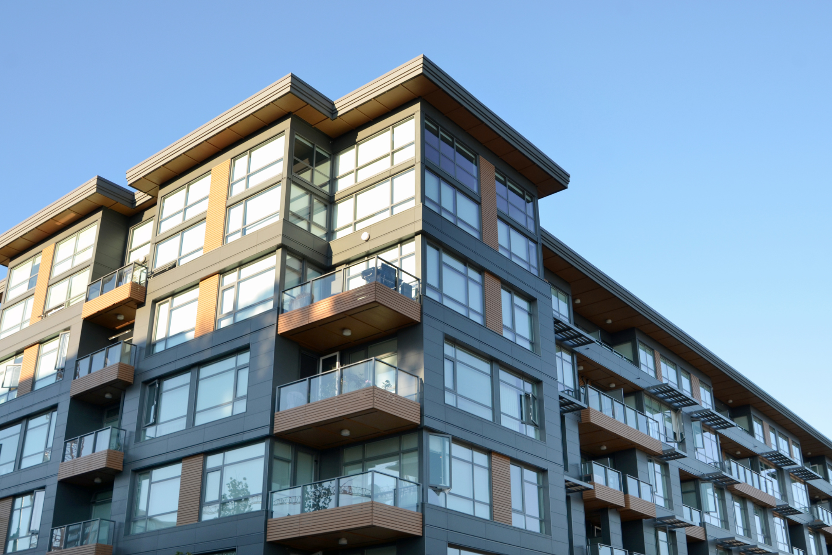 Modern multi-story apartment building with large windows, balconies, and wood accents under a clear blue sky.