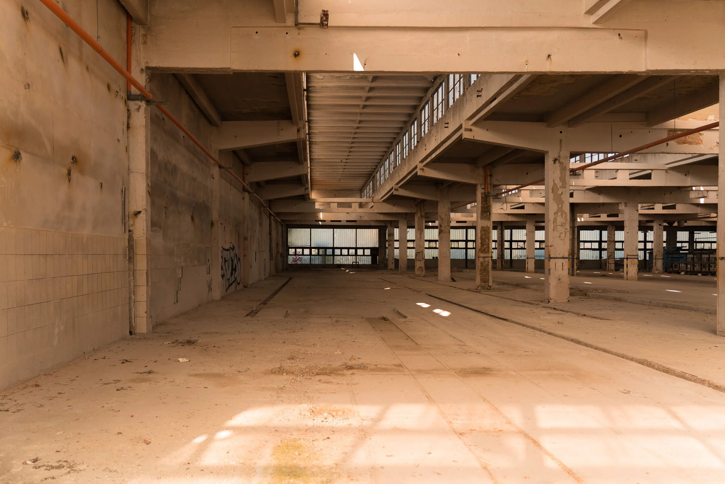 Empty, abandoned indoor space with exposed concrete walls, floors, and ceiling. Support beams and columns are visible, with windows along the upper walls letting in natural light. The area appears to be a vacant industrial or commercial building.