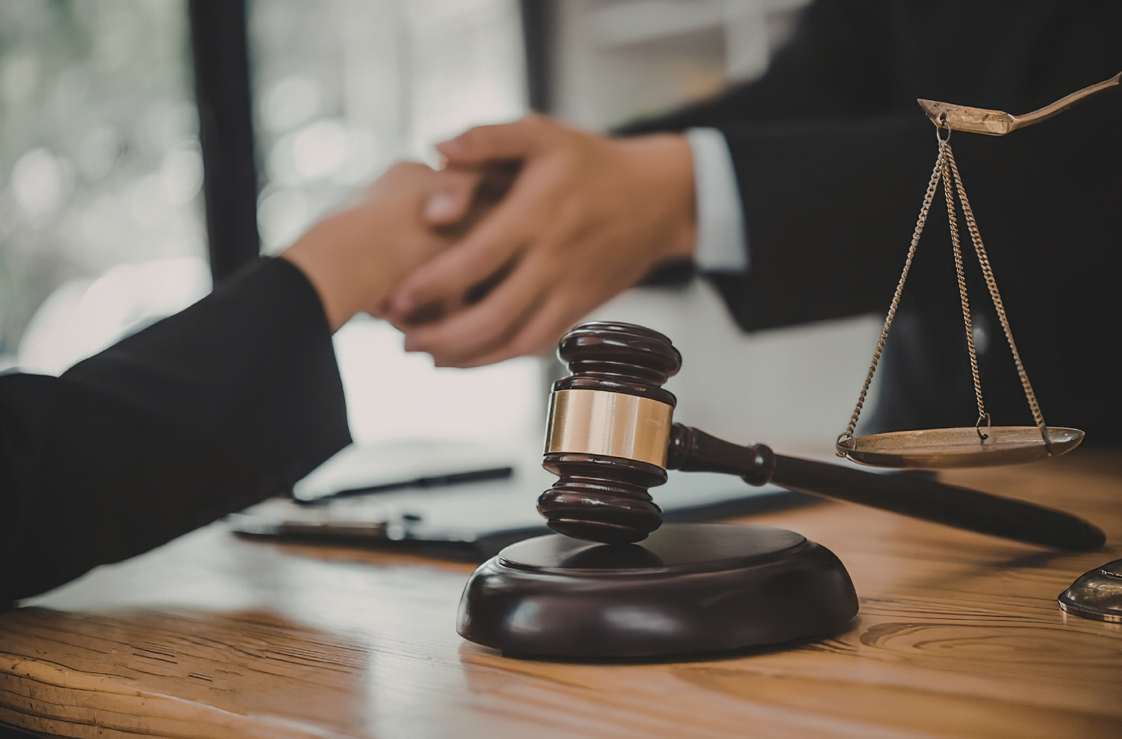 A judge's gavel on a wooden desk with scales of justice in the background, and two people shaking hands, one in a black suit.