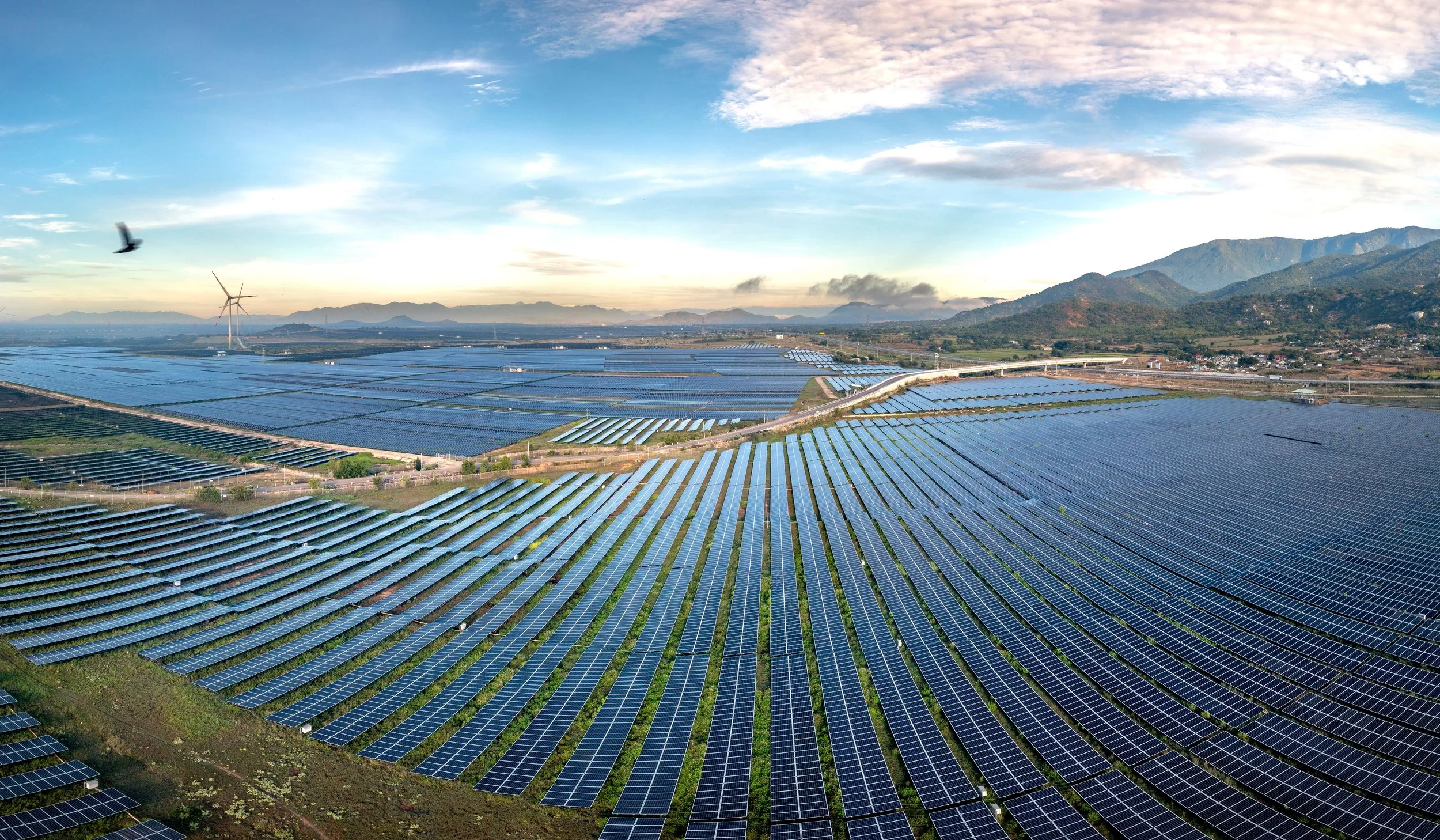 A large solar farm with rows of solar panels stretching across a valley near mountains in the distance. The sky is partly cloudy with a bird flying overhead and a wind turbine visible in the background.