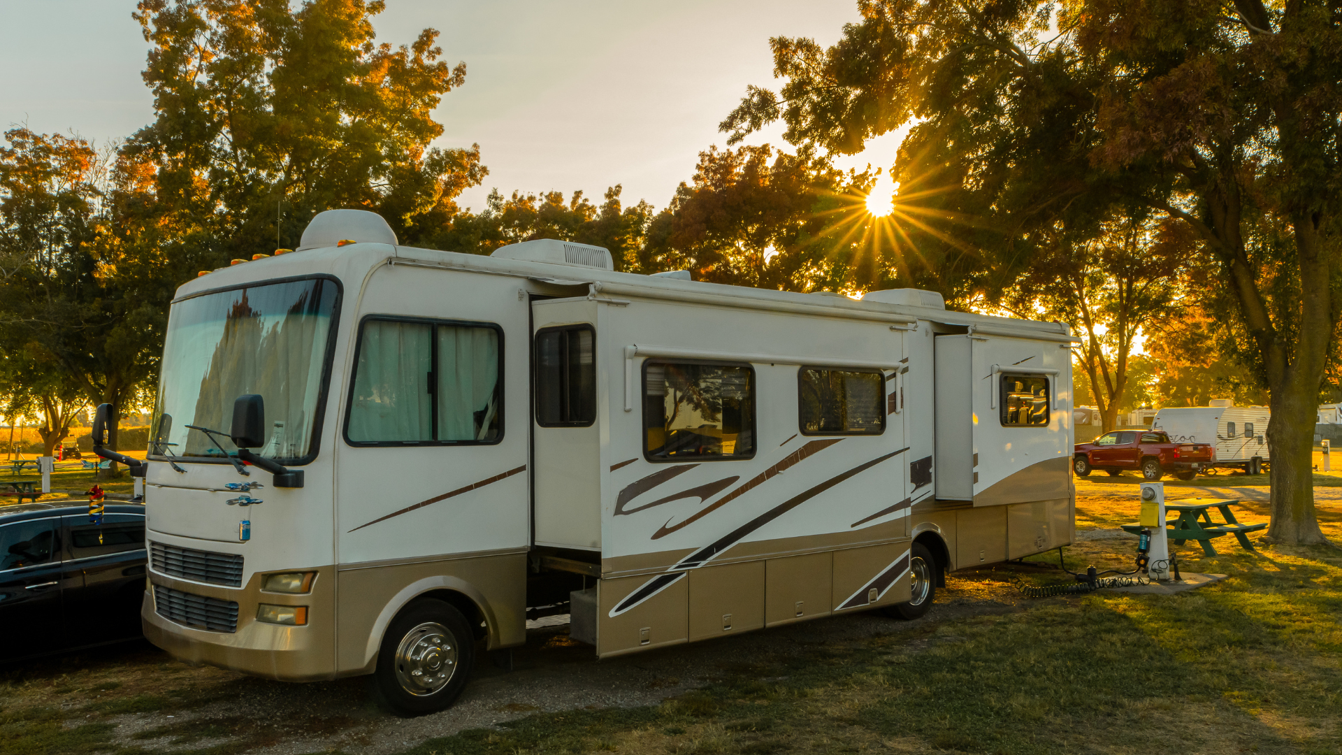 A white and beige RV parked in a campground during sunset with trees, other RVs, cars, picnic tables, and electrical hookups visible.