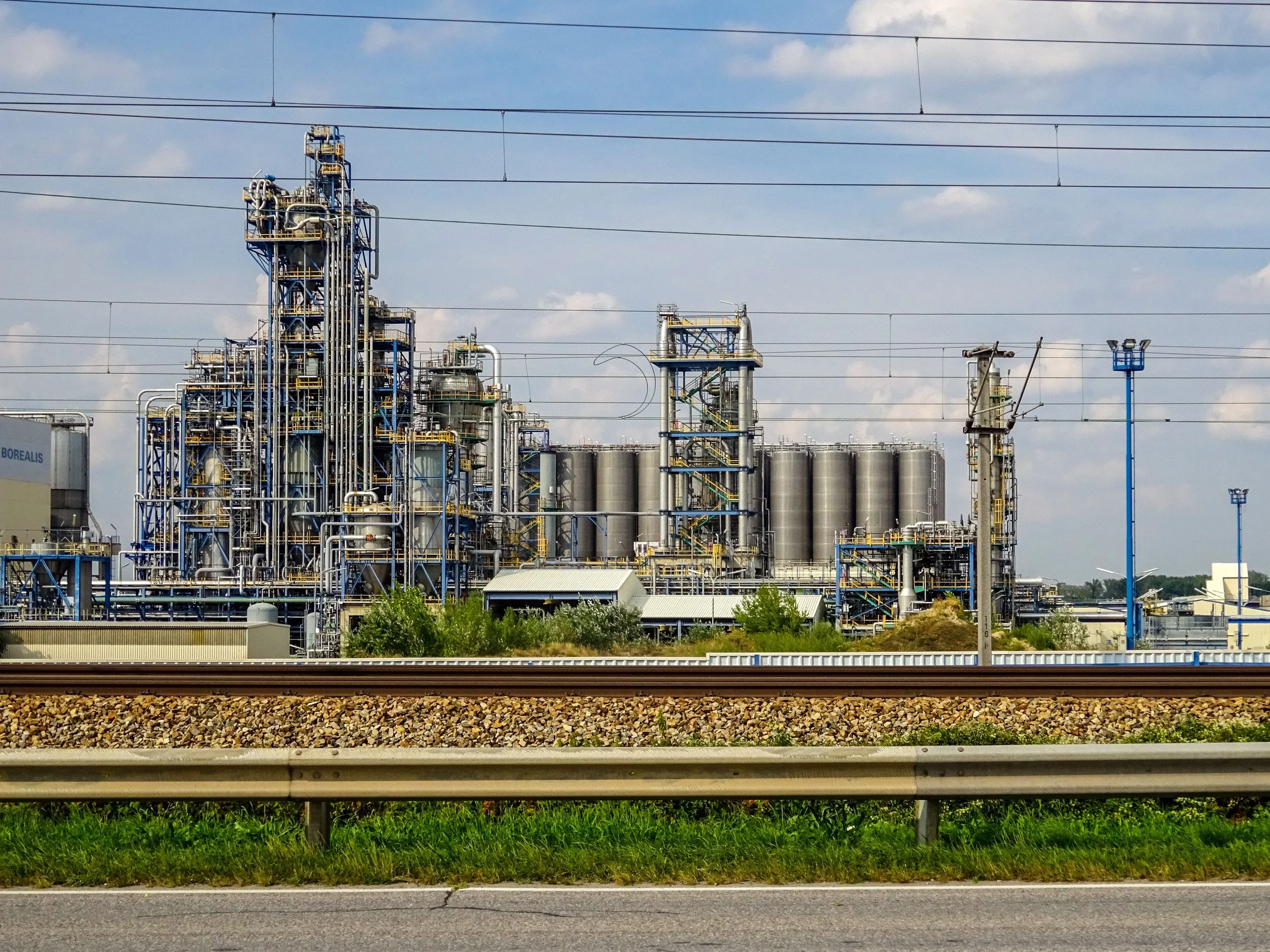 Industrial oil refinery with complex pipelines, storage tanks, and processing units under a partly cloudy sky, viewed from alongside a road with a guardrail in the foreground.