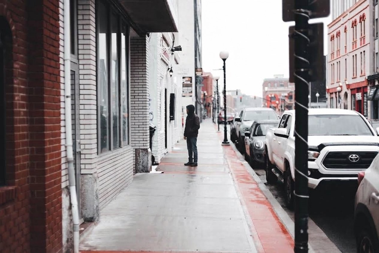 A person stands alone on a city sidewalk next to a row of parked cars, with red and gray buildings in the background on a cloudy day.