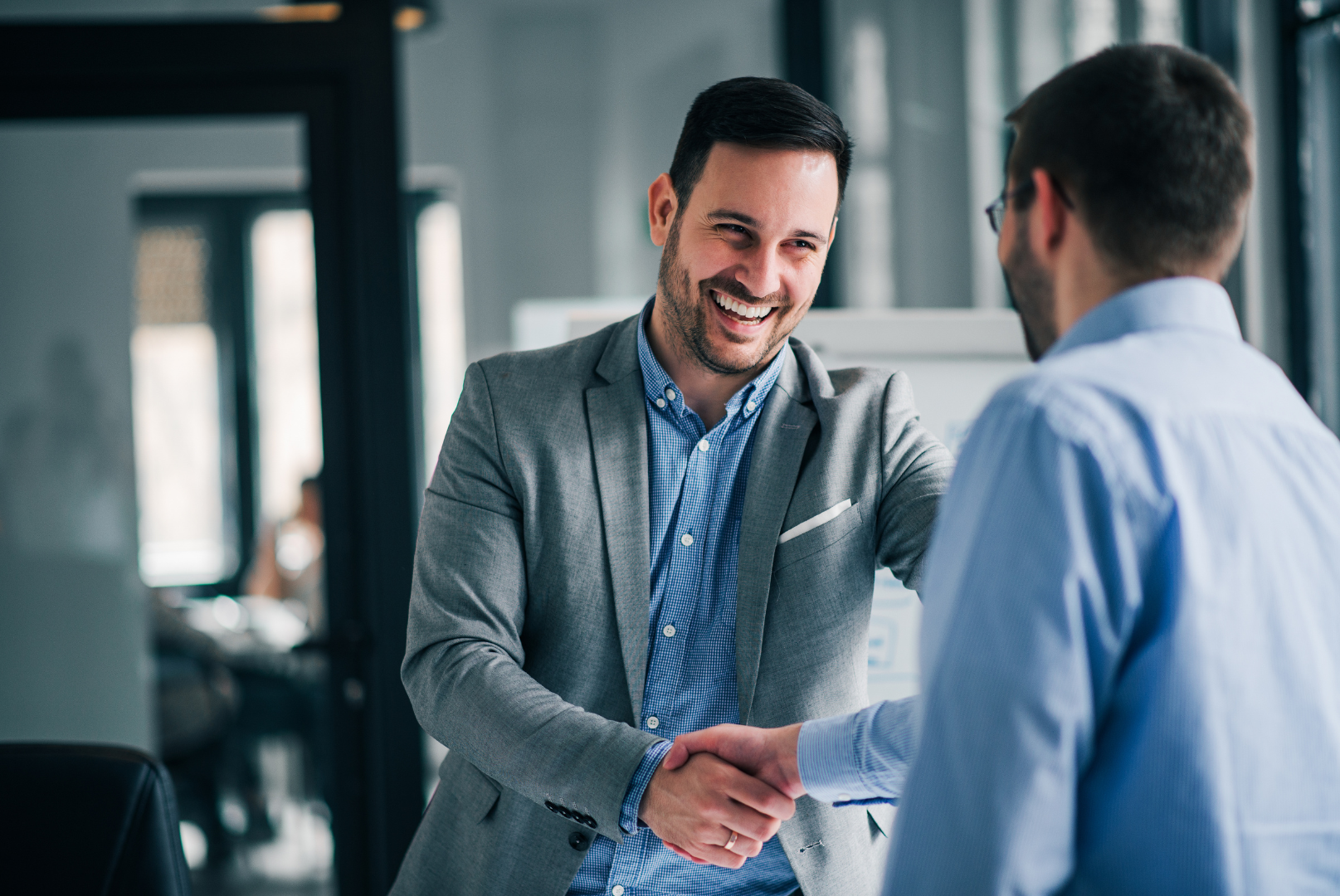 Two men in business attire smiling and shaking hands in a modern office.