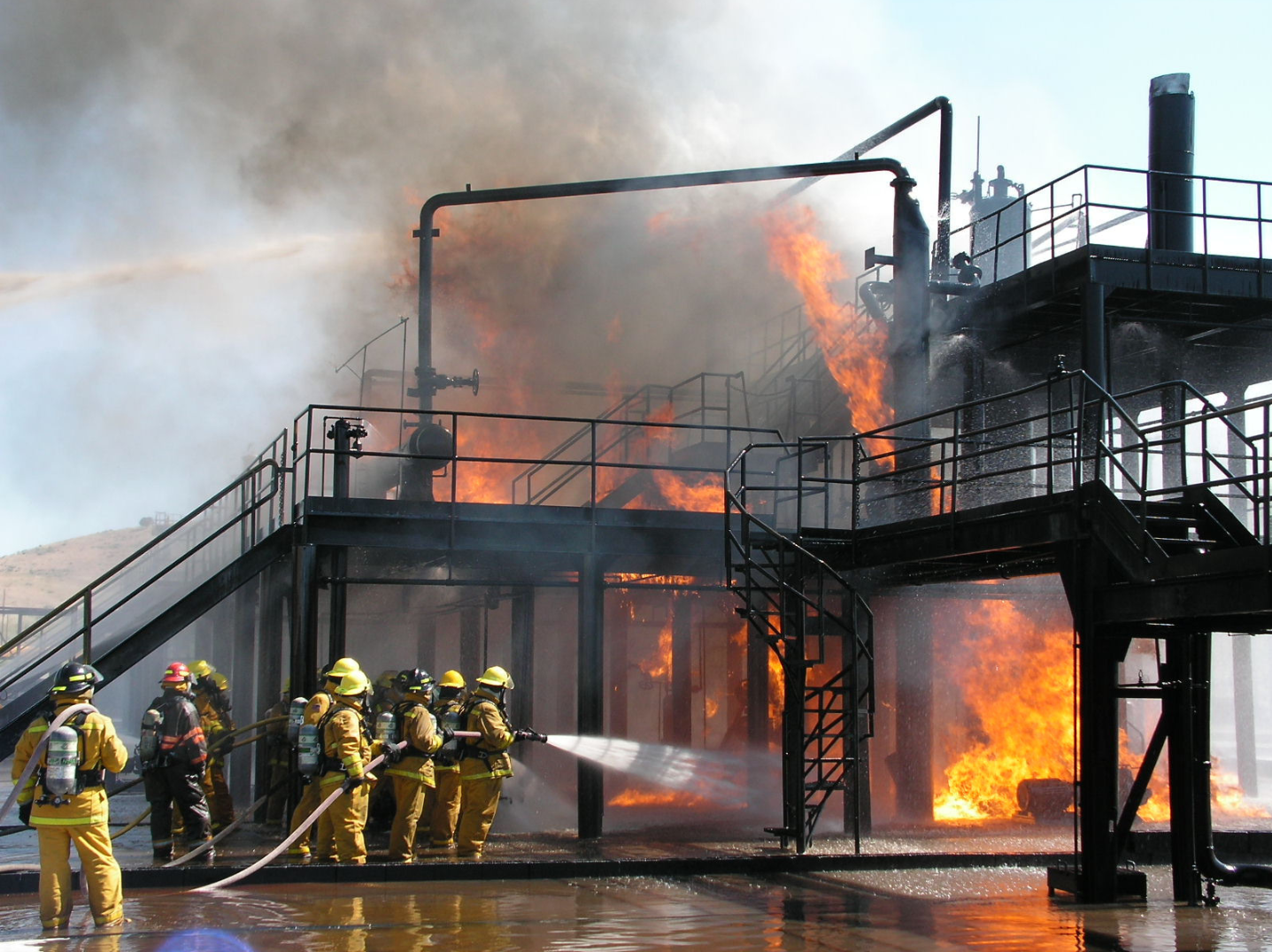 Firefighters battling a large fire on an industrial oil platform, with flames and thick black smoke rising, and firefighters spraying water to extinguish the blaze.