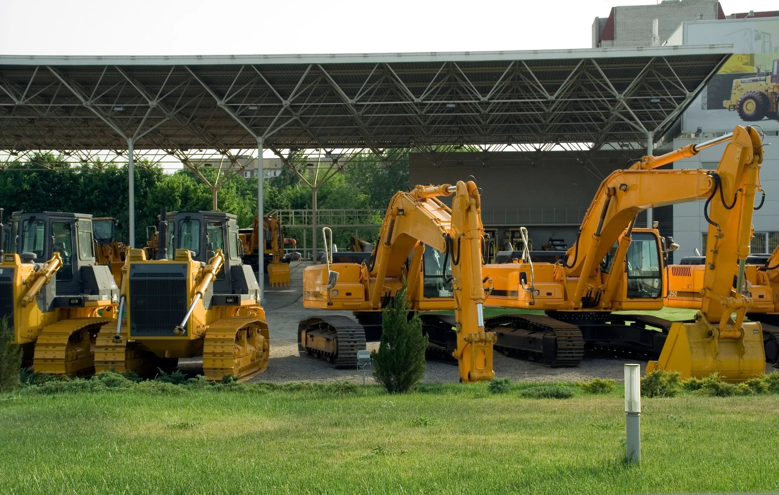 Collection of yellow construction vehicles, including excavators and bulldozers, parked under a metal canopy with greenery in the background.