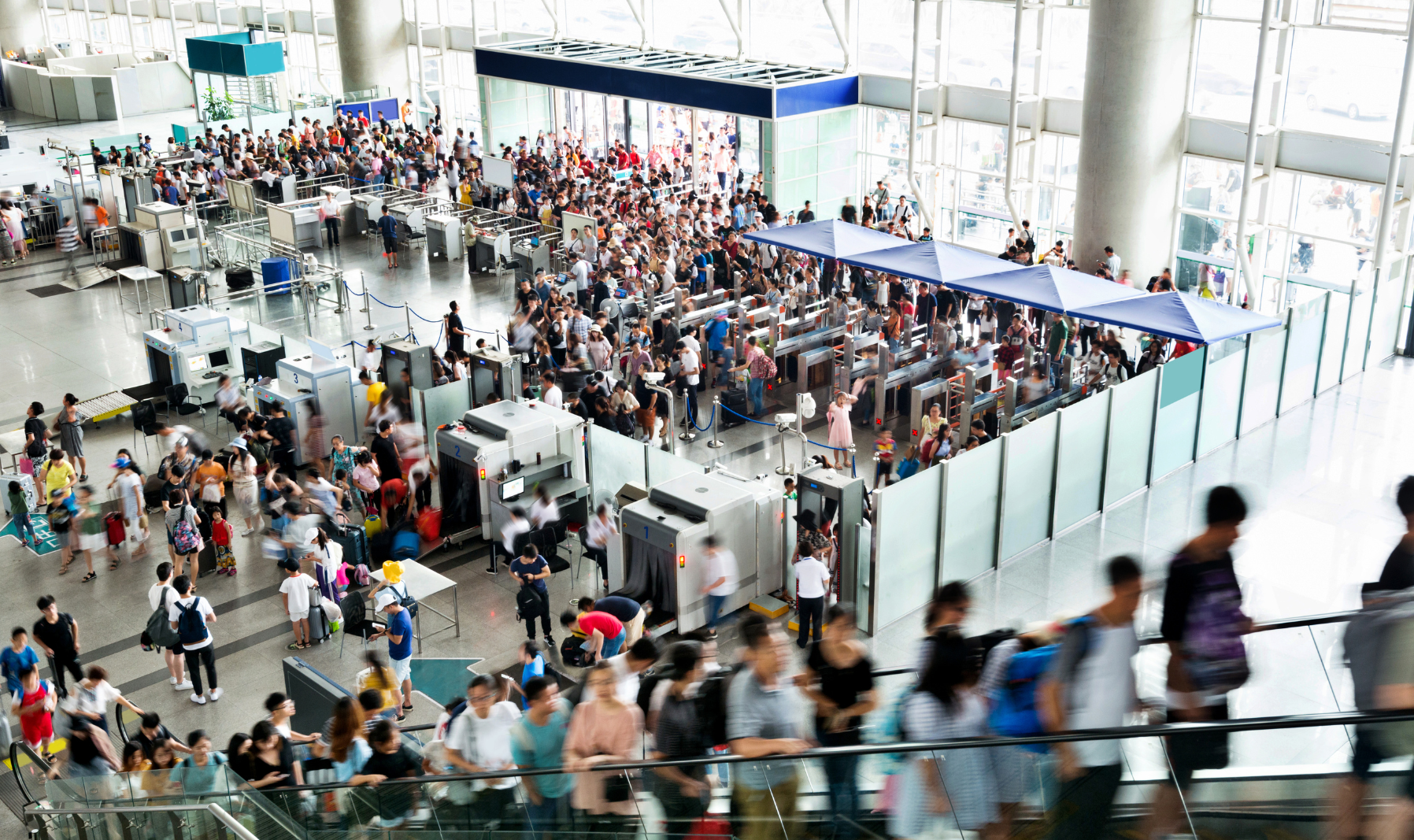 Crowded airport terminal with people waiting in line at security screening, many with luggage, large glass windows, and metal detector stations.