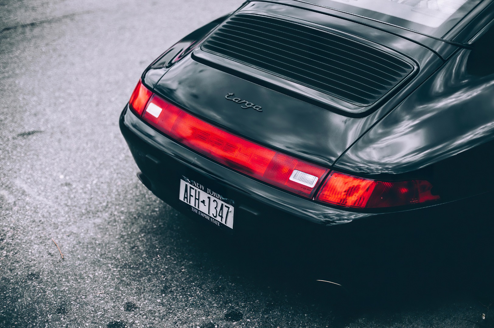 Rear view of a vintage black Porsche 911 Targa with a New York license plate, parked on a textured asphalt surface.