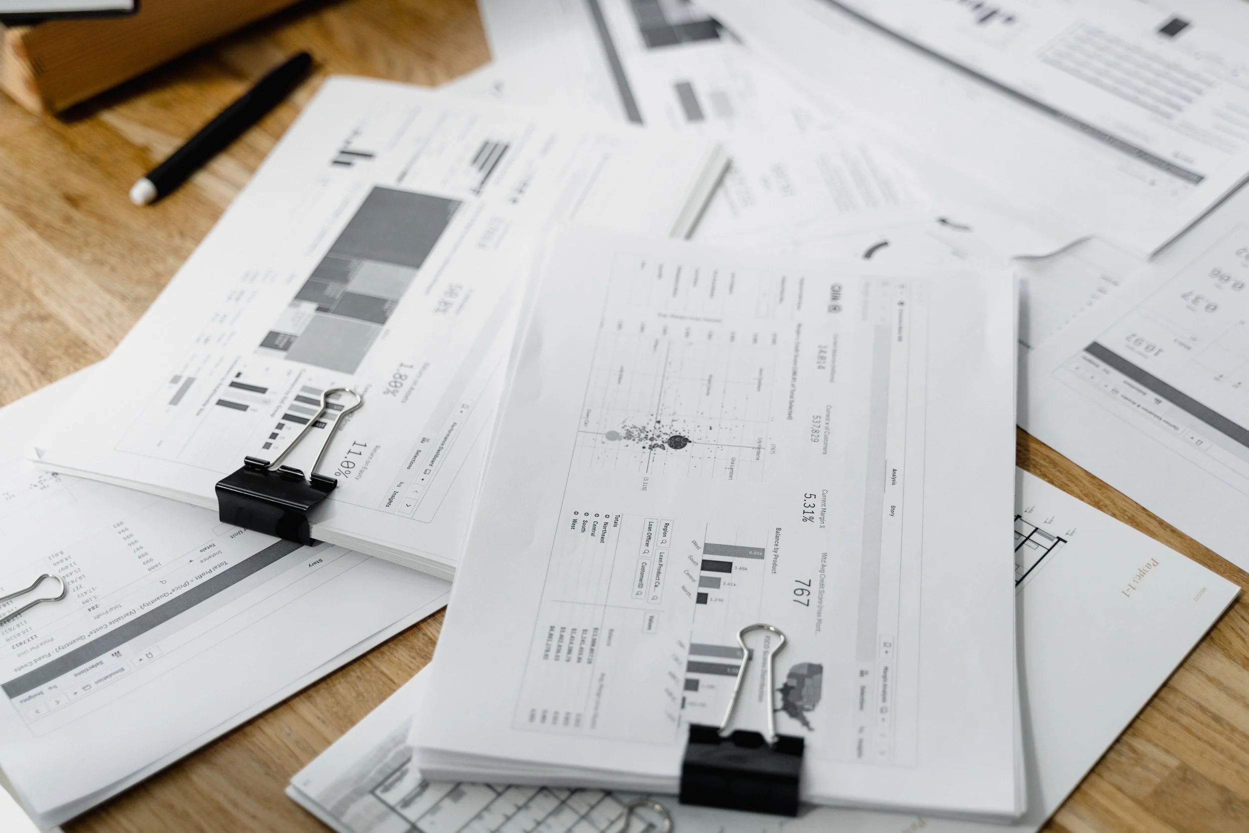 Disorganized papers with charts and graphs scattered on a wooden table, held together by black binder clips. A black marker and a wooden box are also visible.