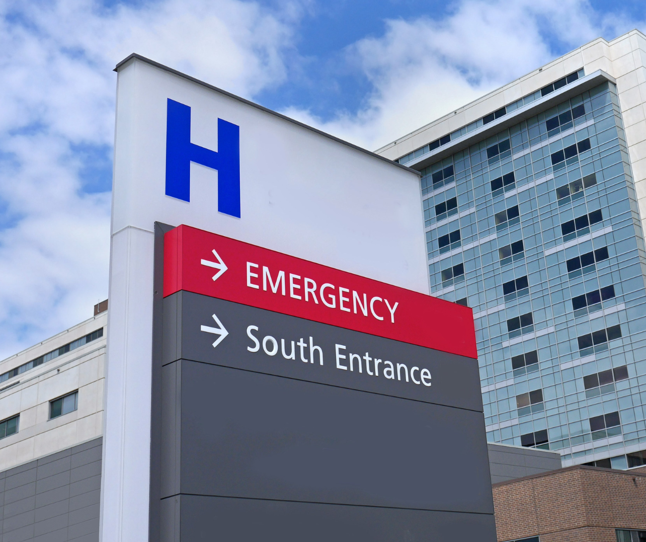Hospital sign with a large letter 'H' at the top, arrow pointing right for Emergency, and arrow pointing right for South Entrance, in front of modern buildings and a partly cloudy sky.