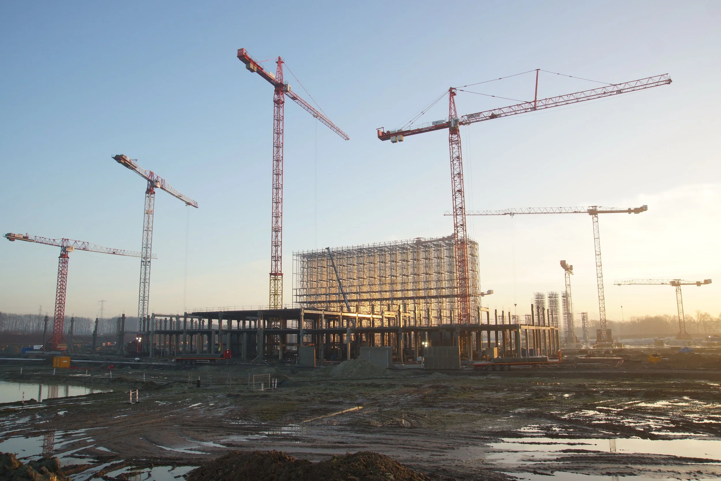 Construction site at sunset with multiple red tower cranes and scaffolding for a building under construction, muddy ground with puddles.