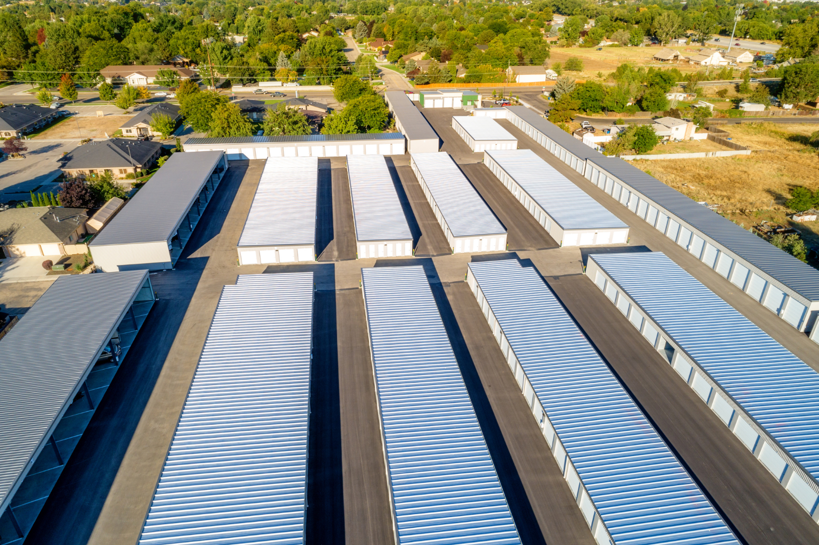 Aerial view of a large self-storage facility with multiple rows of white storage units and covered parking areas, surrounded by a residential neighborhood and green trees.