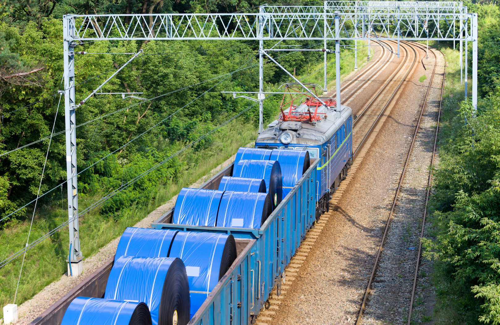An electric train carrying large blue coils on freight cars traveling on a railway track surrounded by green trees.