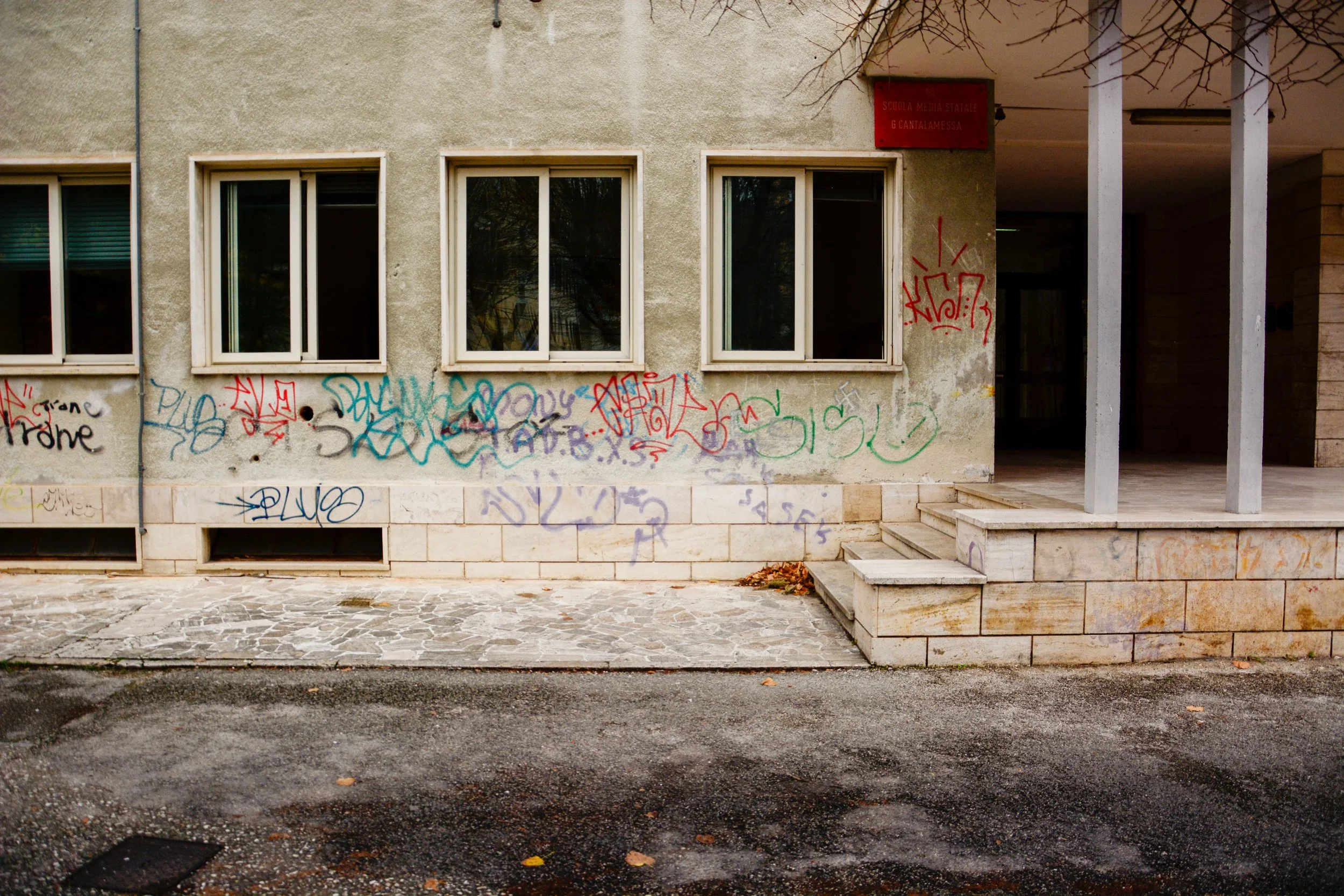 The exterior wall of a building with three windows, graffiti on the wall, a small staircase leading to a covered entrance, and part of a tree with bare branches.