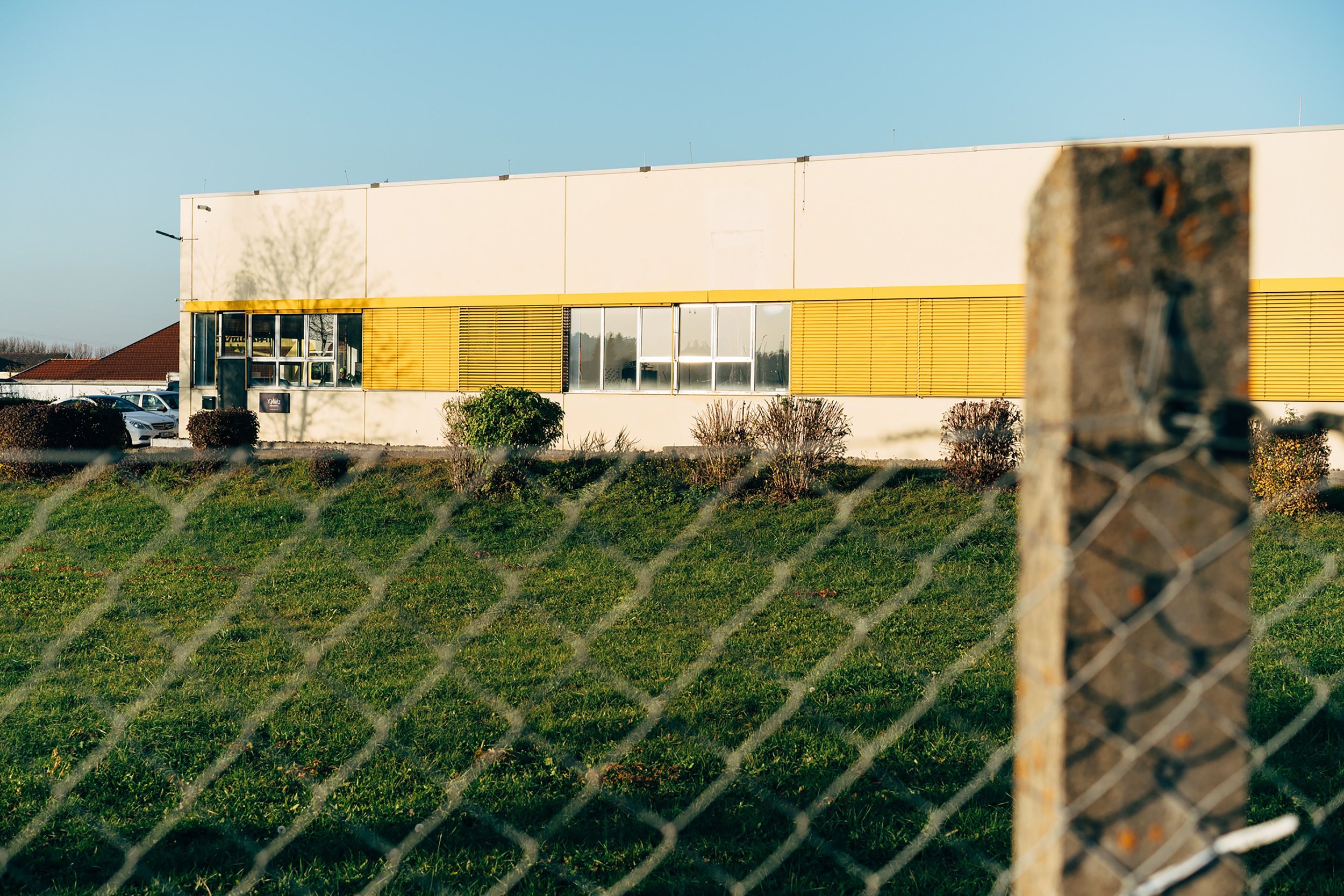 A white building with yellow accents and large windows, surrounded by cars and greenery, seen through a chain-link fence and blurred wooden post in the foreground that surrounds the building.