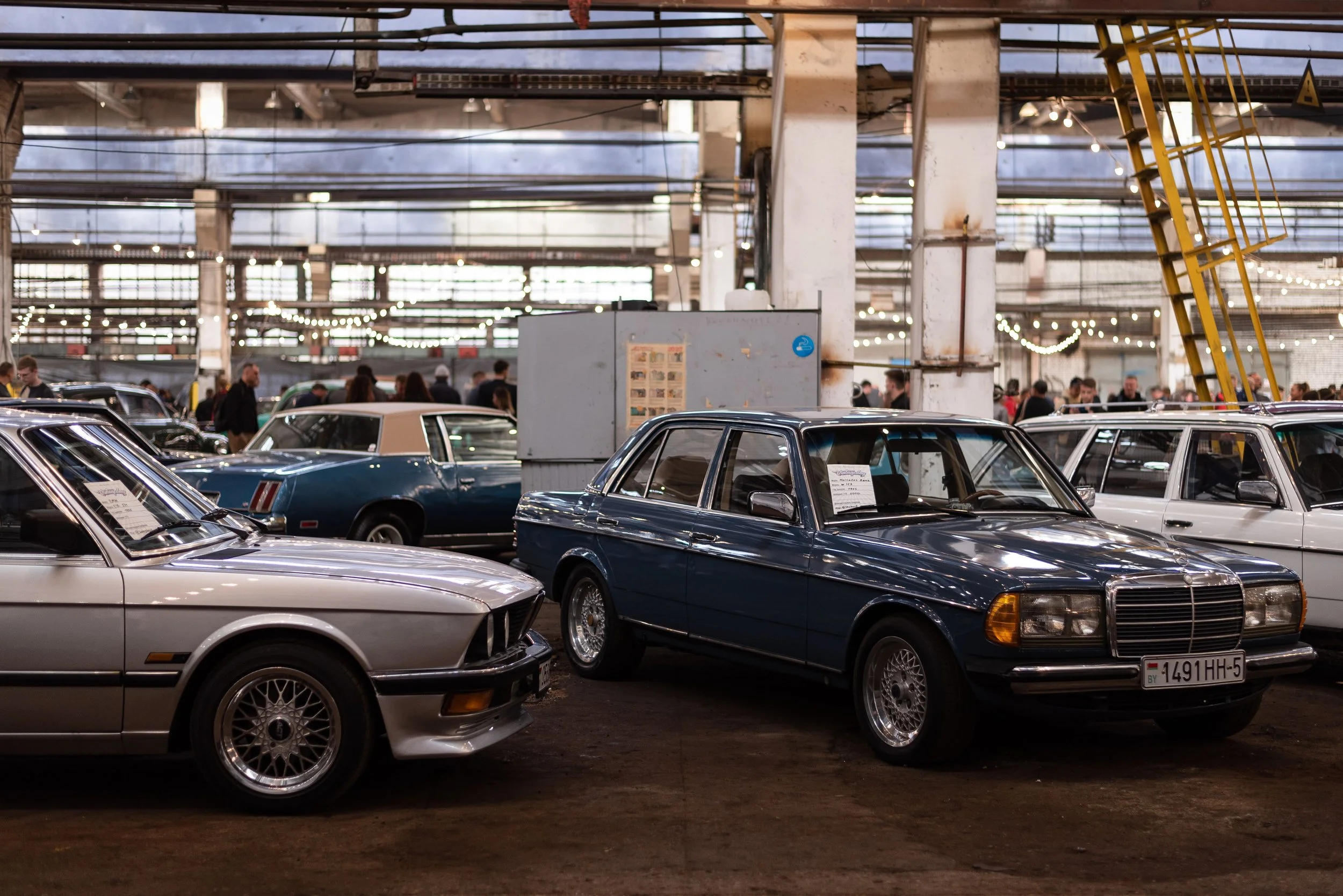 Vintage cars on display inside a large industrial building with string lights and people walking around.