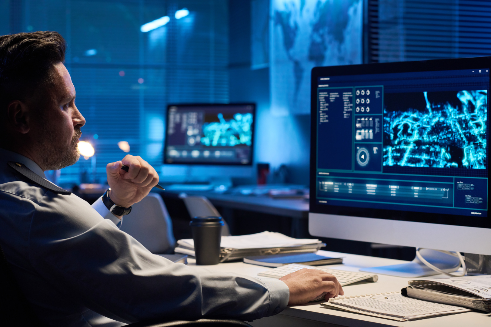A man working at a desk with multiple computer monitors displaying digital maps and surveillance camera data in a dark office.