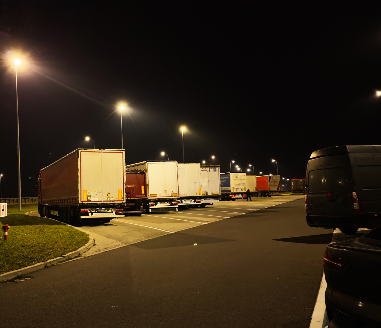 Nighttime scene of semi trucks parked in a well-lit parking lot, with streetlights illuminating the area and a few pedestrians walking in the distance.