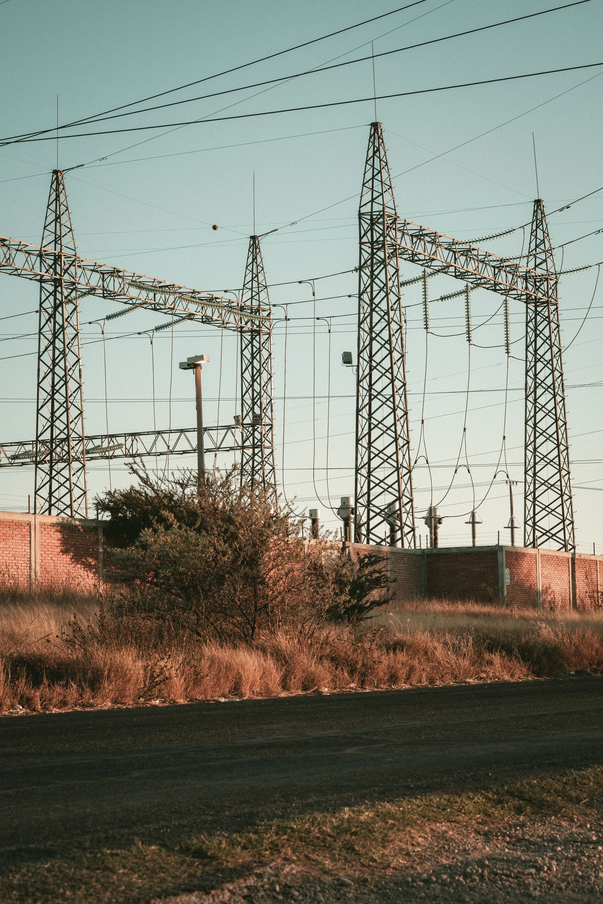 Power lines and electrical towers against a clear sky, with a fence, streetlight, and grassy area in the foreground.