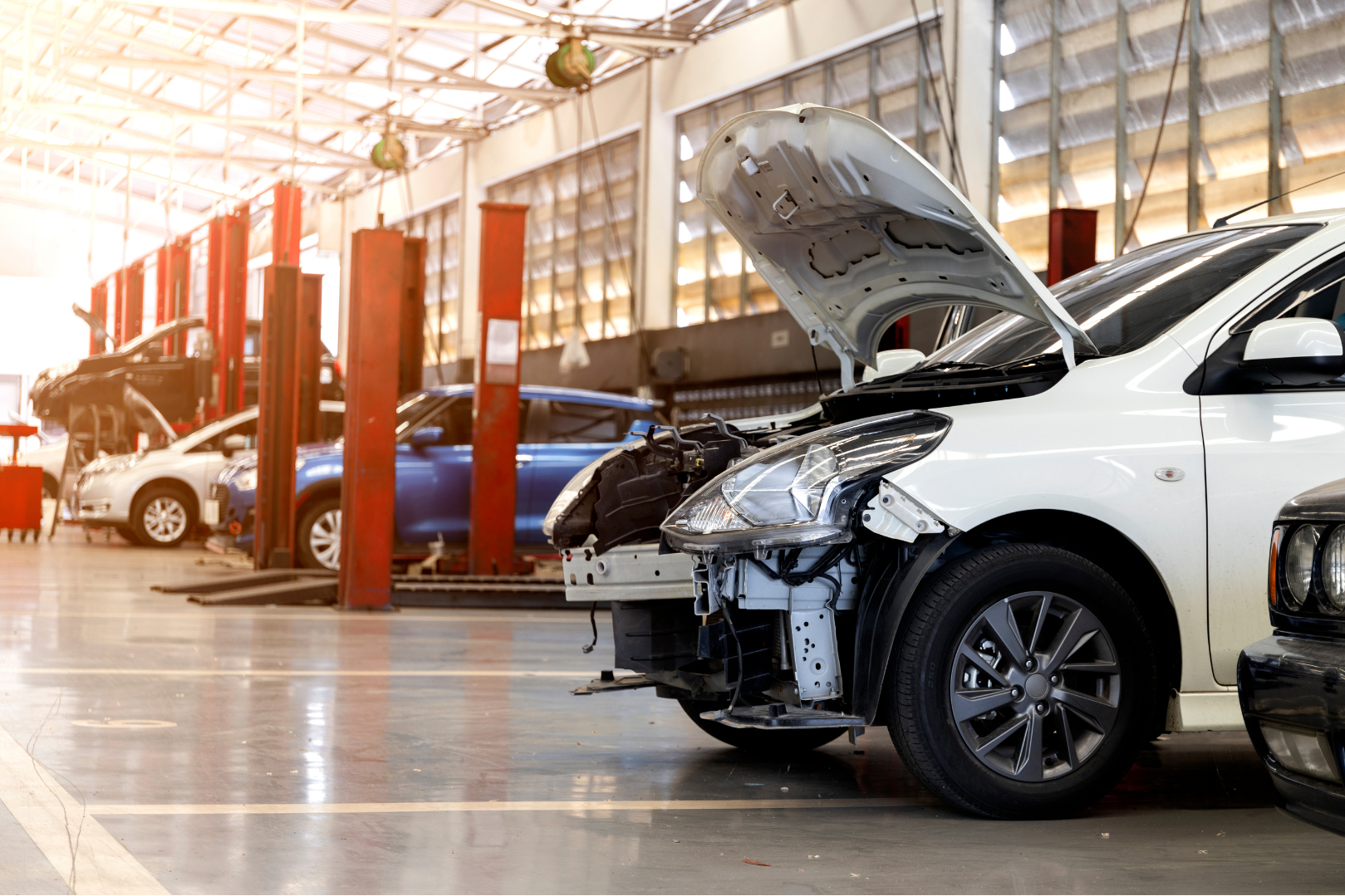Indoor car repair or body shop with multiple cars, including a white car with front damage, in a spacious, well-lit workshop with tall garage lifts.