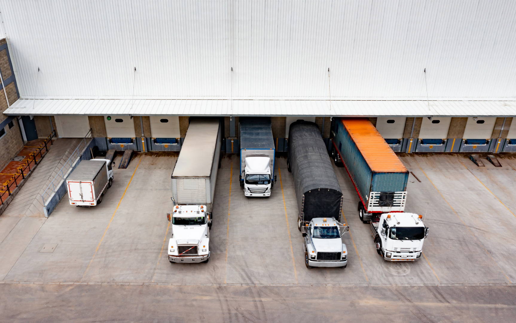 Overhead view of a loading dock area with five parked trucks aligned with dock doors at a warehouse. The trucks vary in size and color, including white, gray, and blue, with some having cargo secured in the trailers.