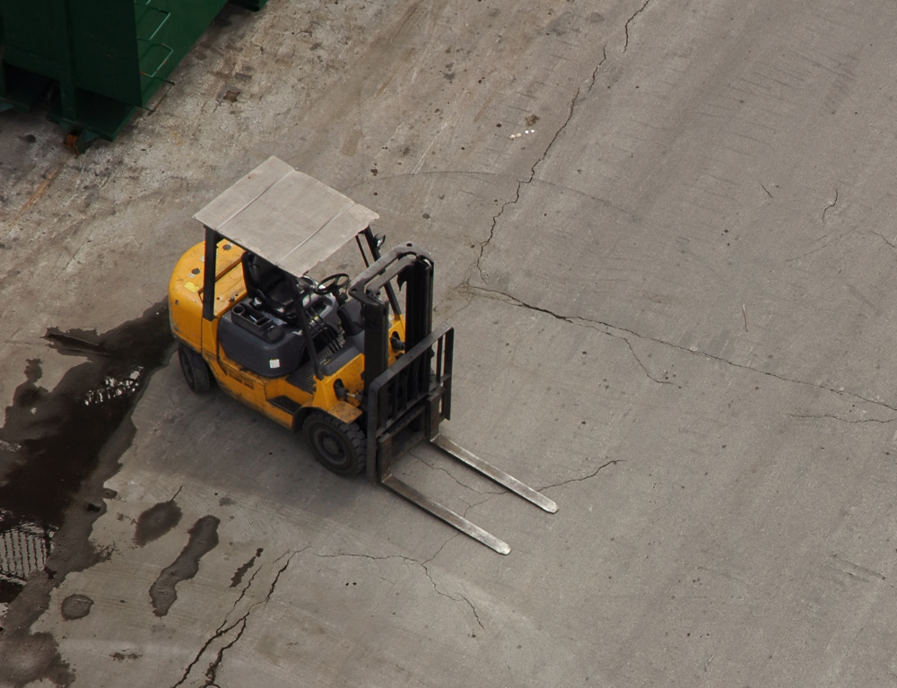 An overhead view of a yellow forklift on a cracked concrete floor, with some puddles of water nearby.