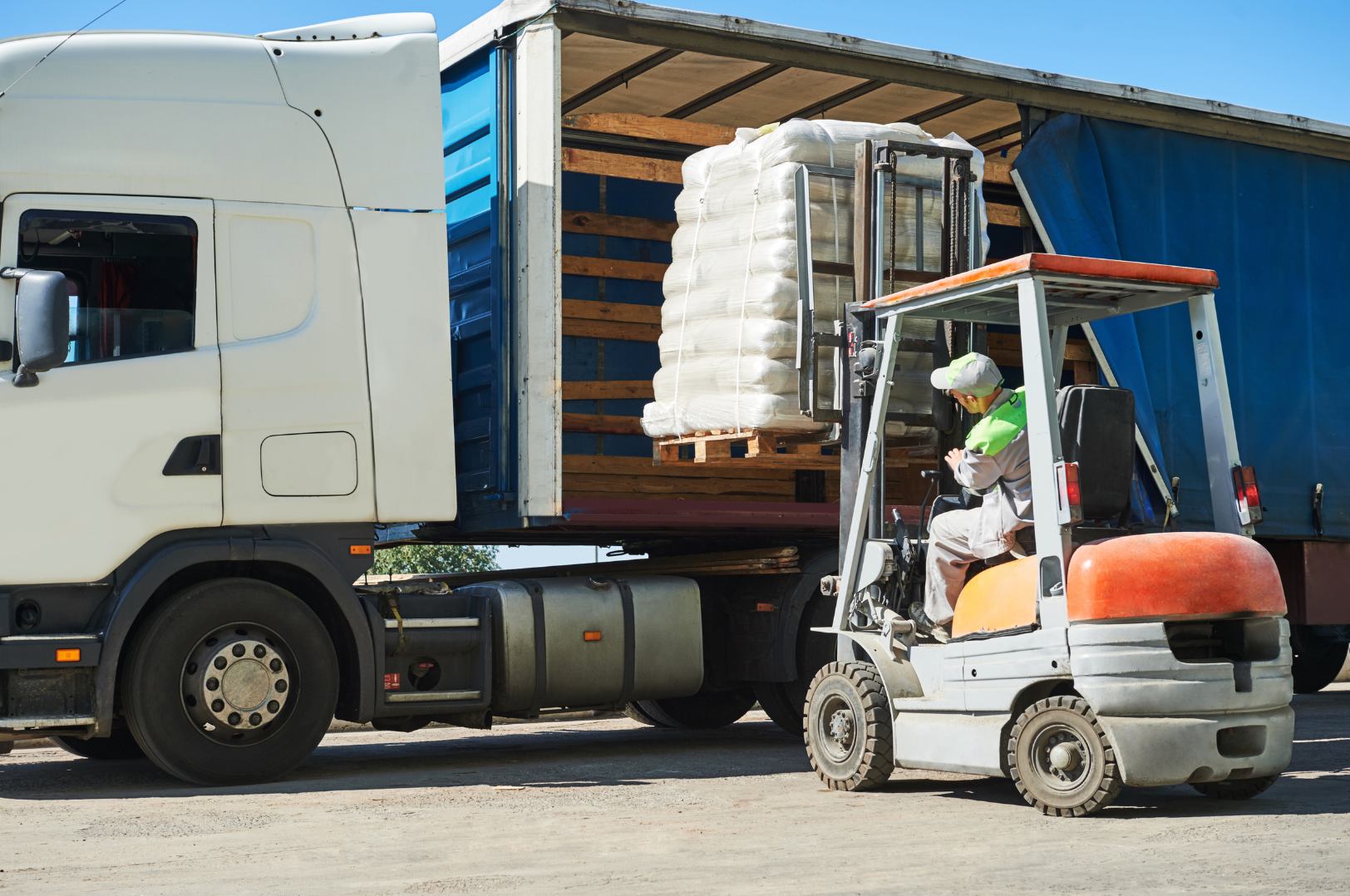 A worker operating a forklift to load a pallet of goods into a large truck. The truck has a half-open back with a blue cover pulled to the side, revealing wooden pallets and large white bags inside.