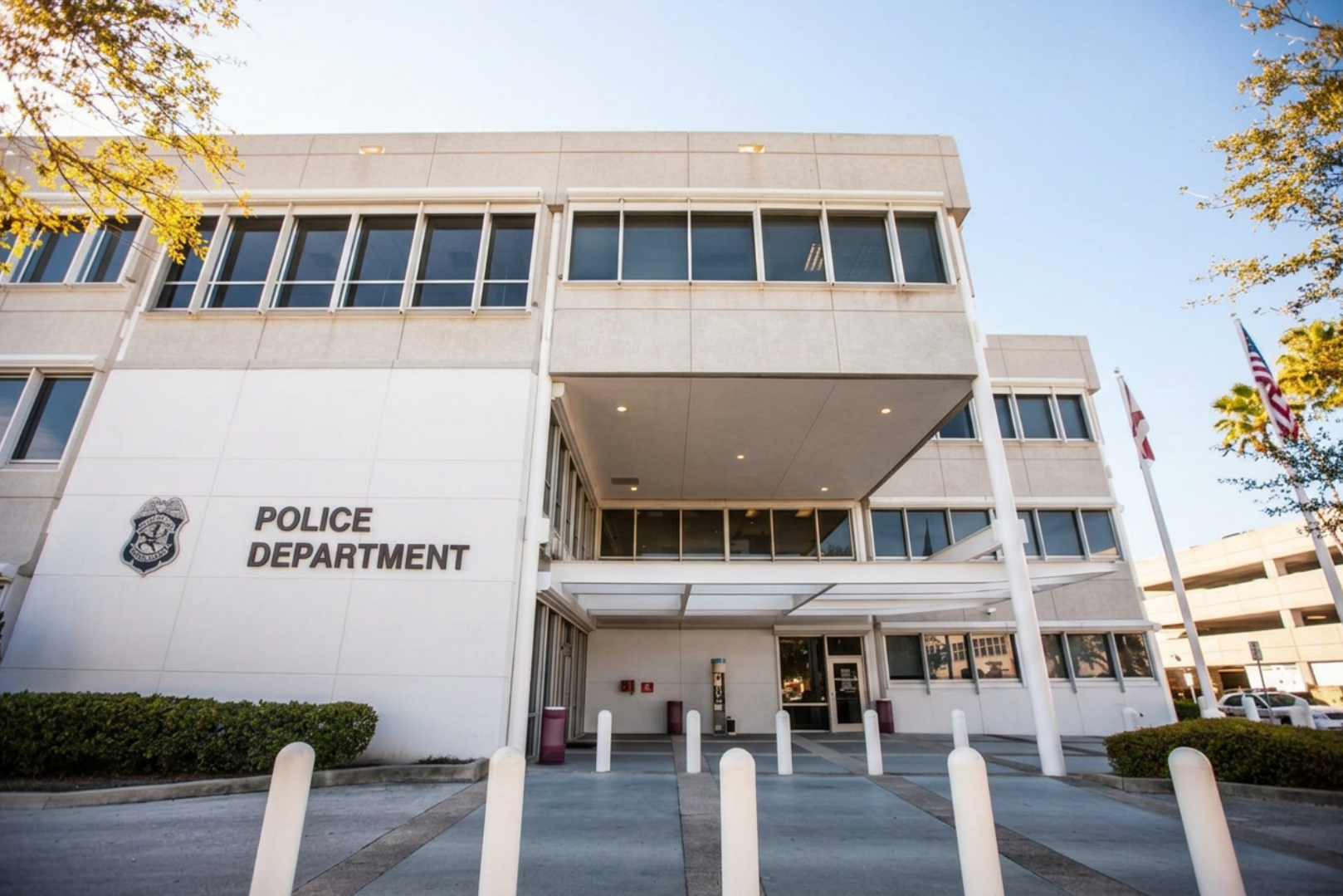 Exterior of a police department building with the sign 'Police Department' and a police badge logo, flags, and a small driveway.