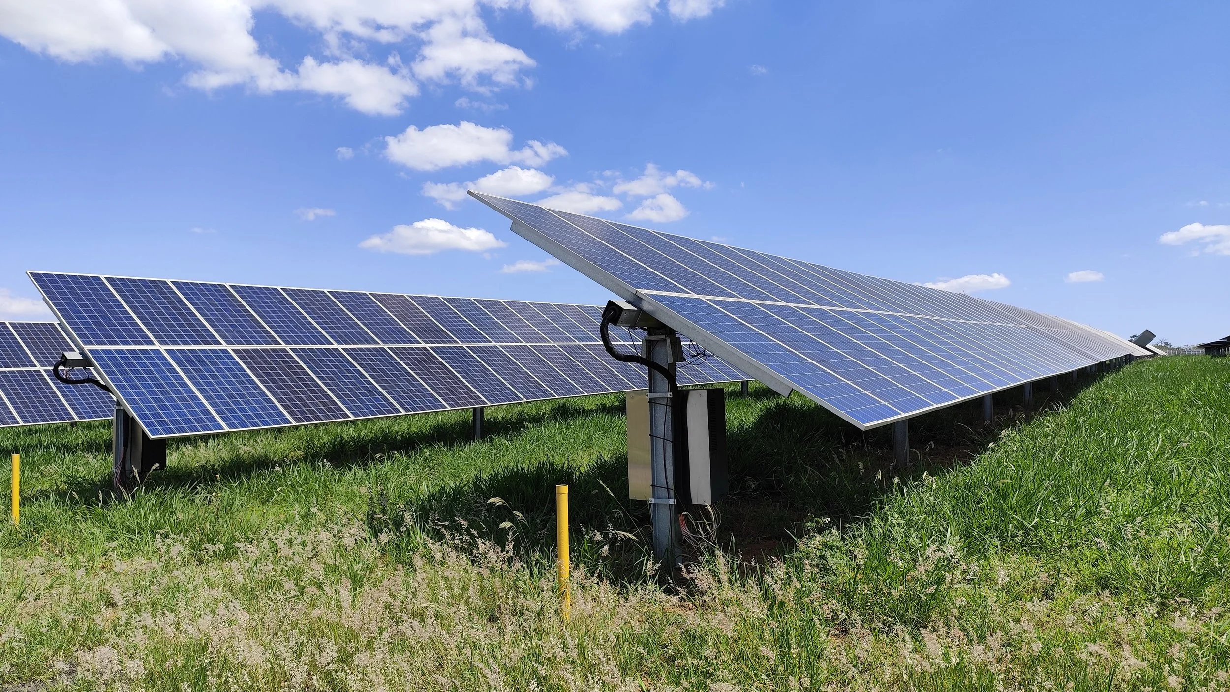 Multiple solar panels installed on grassy land under a blue sky with scattered clouds.