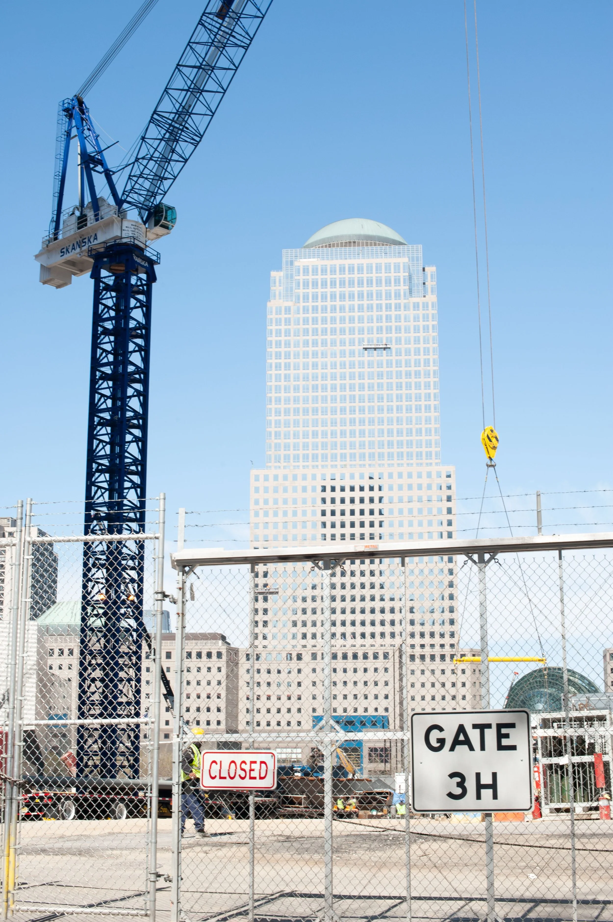 Construction site with a blue crane and a tall building in the background, fenced off with signs that read 'CLOSED' and 'GATE 3H' in front.