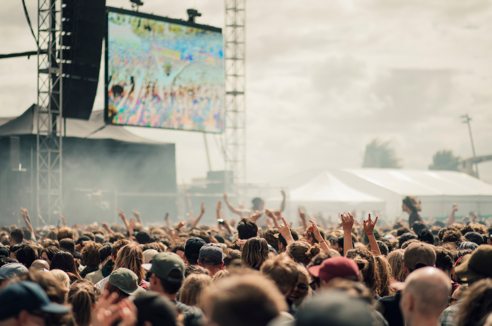 Crowd at outdoor concert with large screen, stage, and tents under cloudy sky.