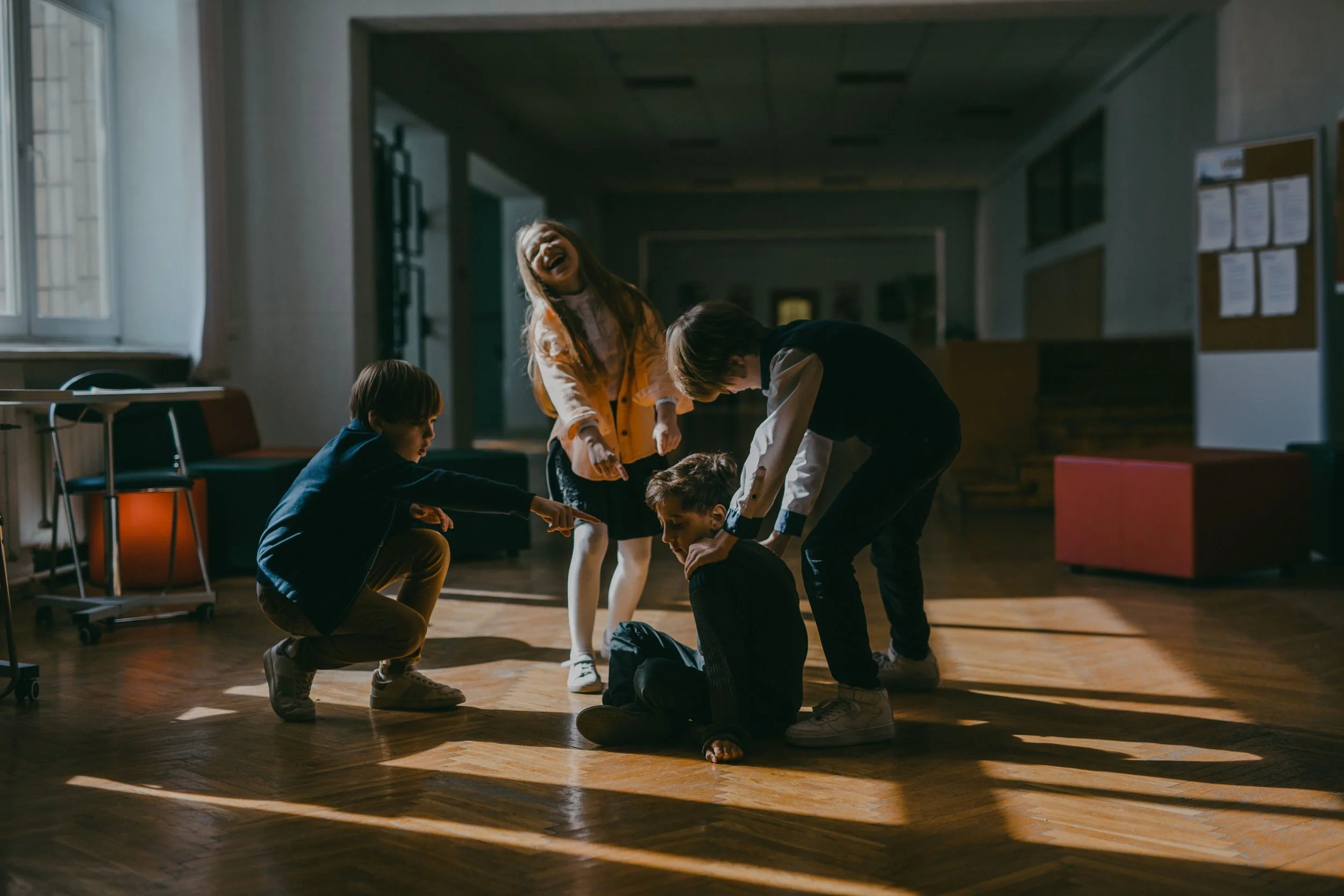 3 school kids surrounding and bullyinh a boy sitting on the ground, one boy and girl point at him laughing, and the third is touching his shoulders.