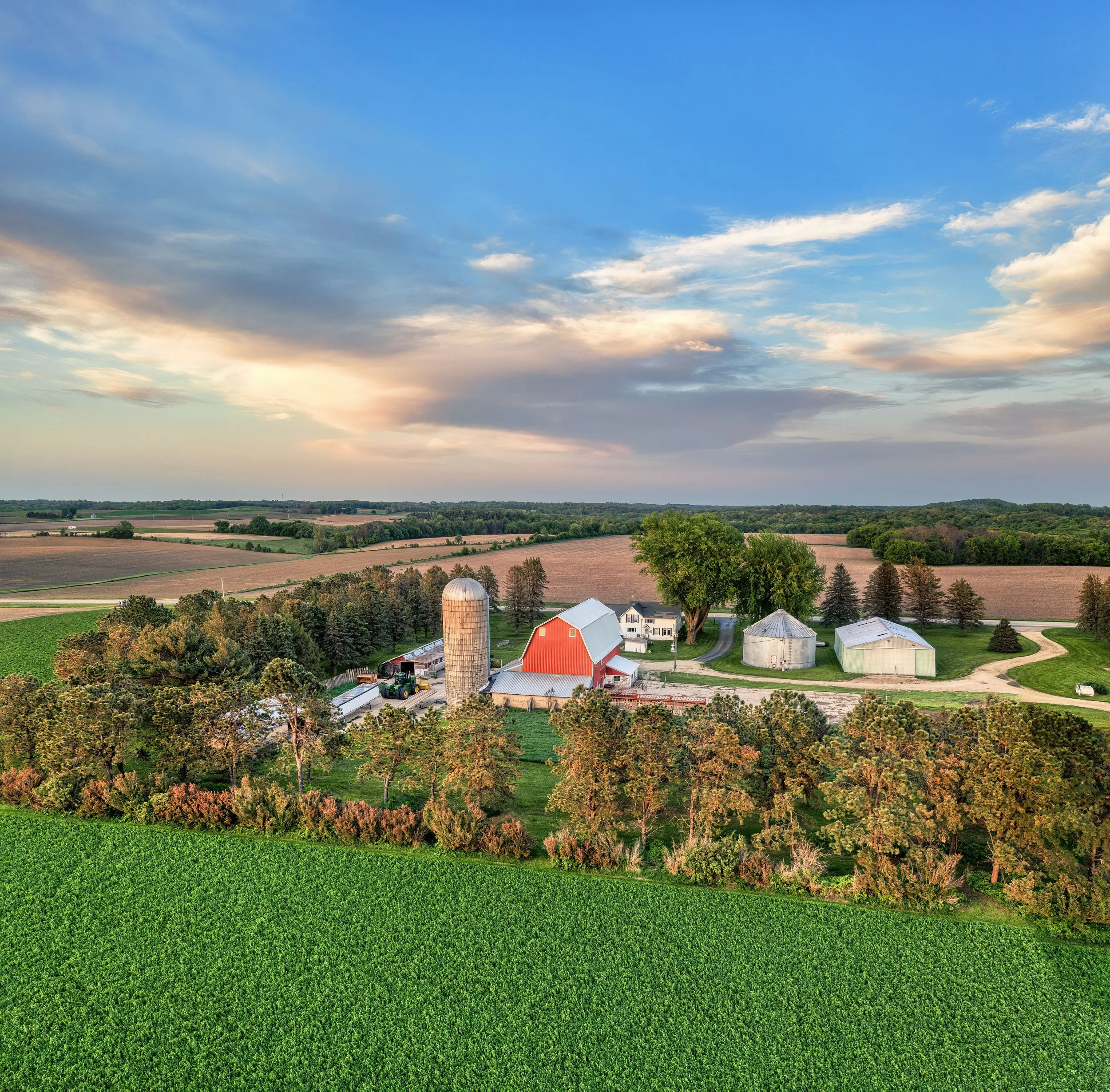 A rural landscape with a farm, barns, silos, and fields under a partly cloudy sky during daytime.