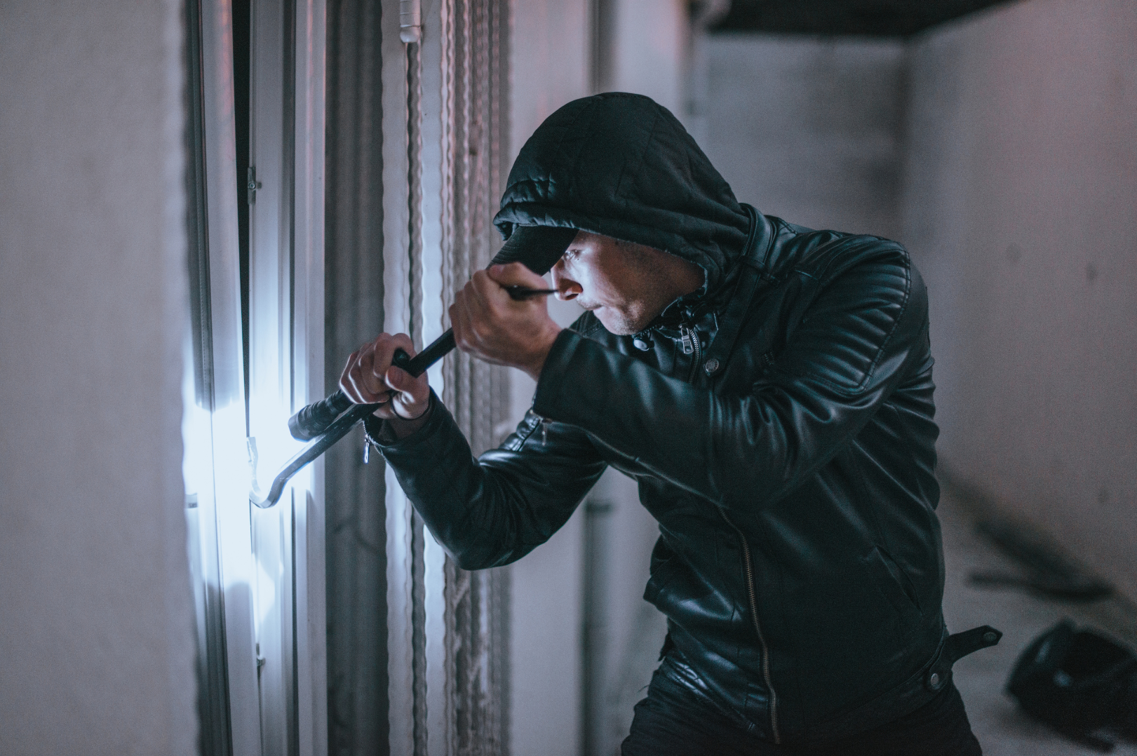 Man wearing a black hoodie and leather jacket attempting to pry open a door with a crowbar.