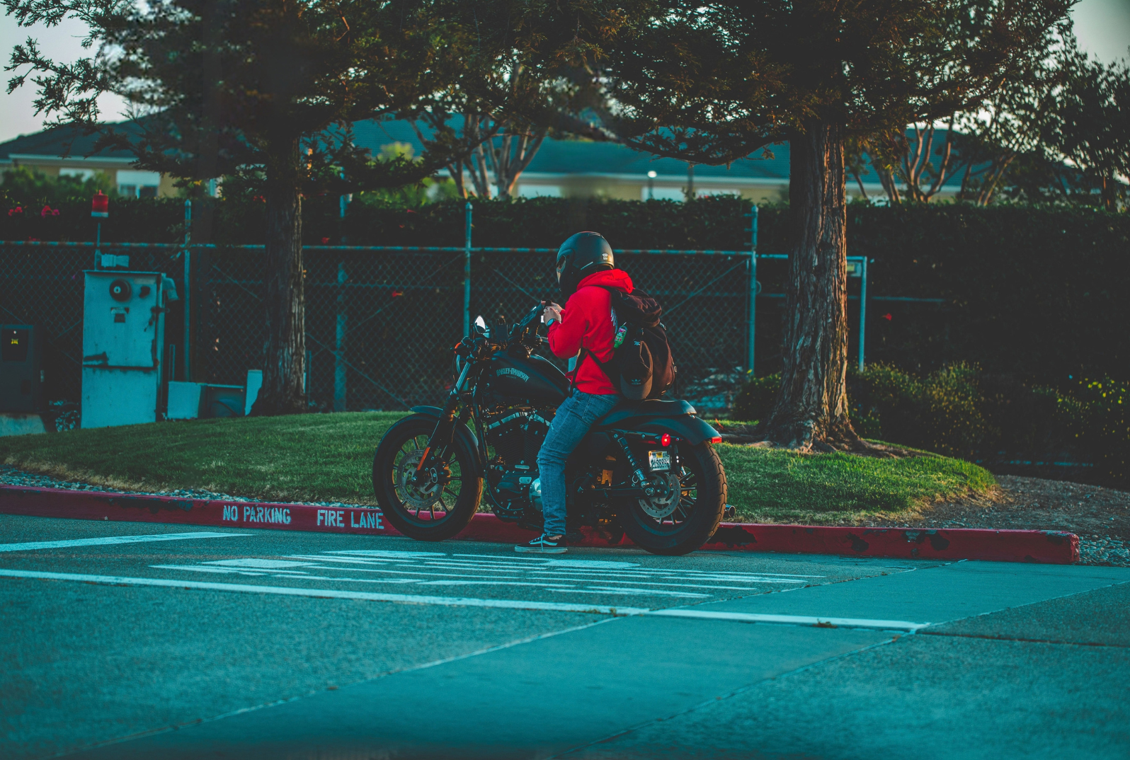 A person in a red hoodie and jeans sitting on a black motorcycle, wearing a black helmet and backpack, parked near a curb with a 'No Parking Fire Lane' sign, next to a large tree and a chain-link fence.