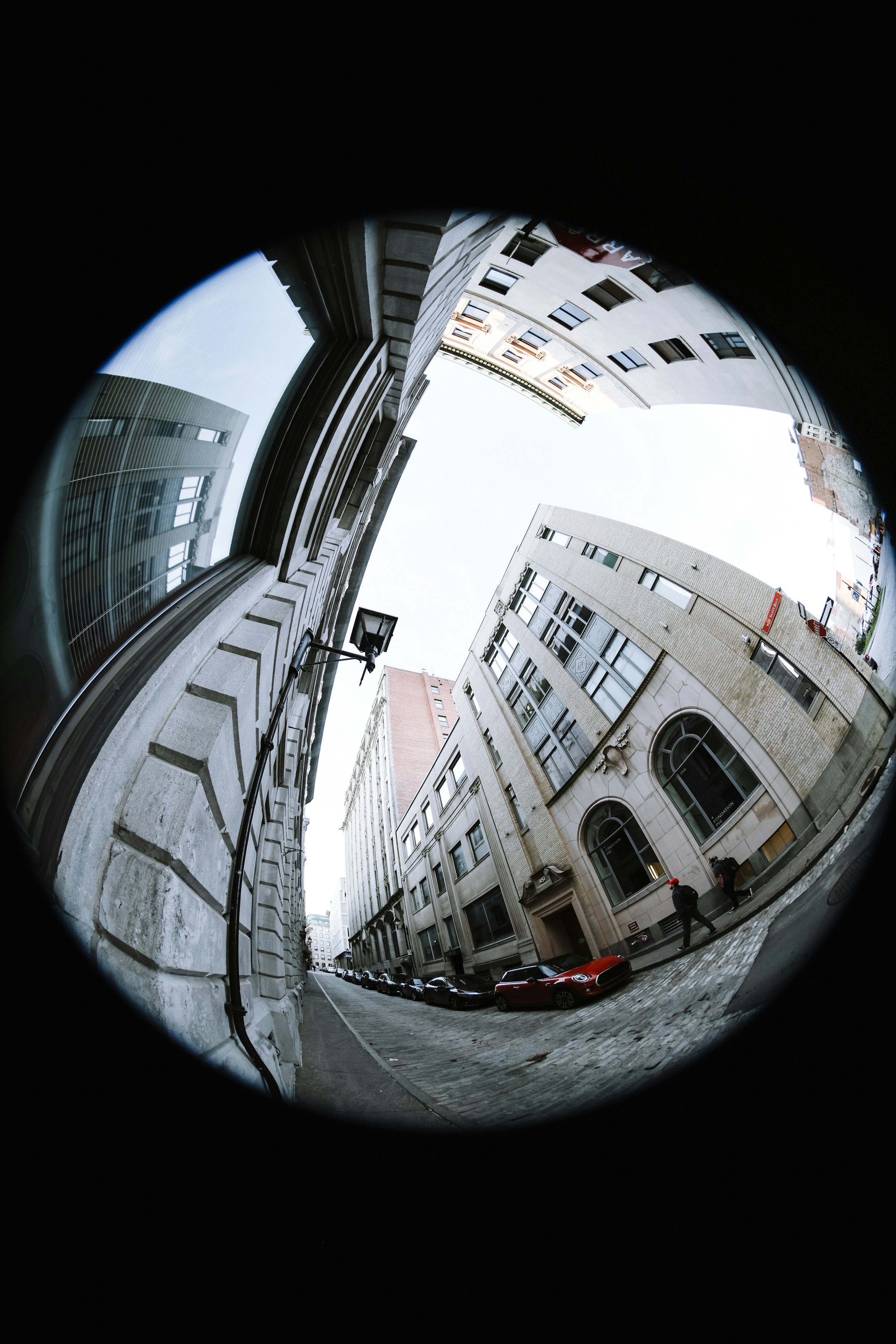Looking up at a city street through a fisheye lens, showing tall buildings with windows, parked cars along the curb, and a few pedestrians walking on the sidewalk.