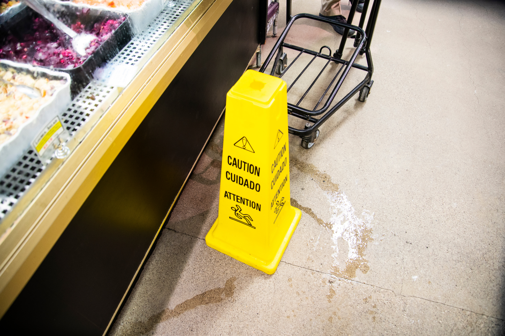 A yellow caution wet floor sign placed on the store floor near the refrigerated section, with a shopping cart beside it.