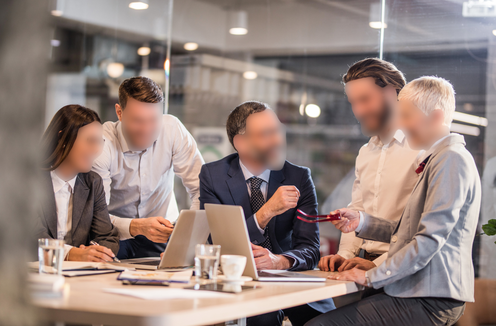 Group of five professionals, four men and one woman, all their face blurred using ai privacy tools, engaged in a discussion around a table with laptops and documents in a modern office setting.