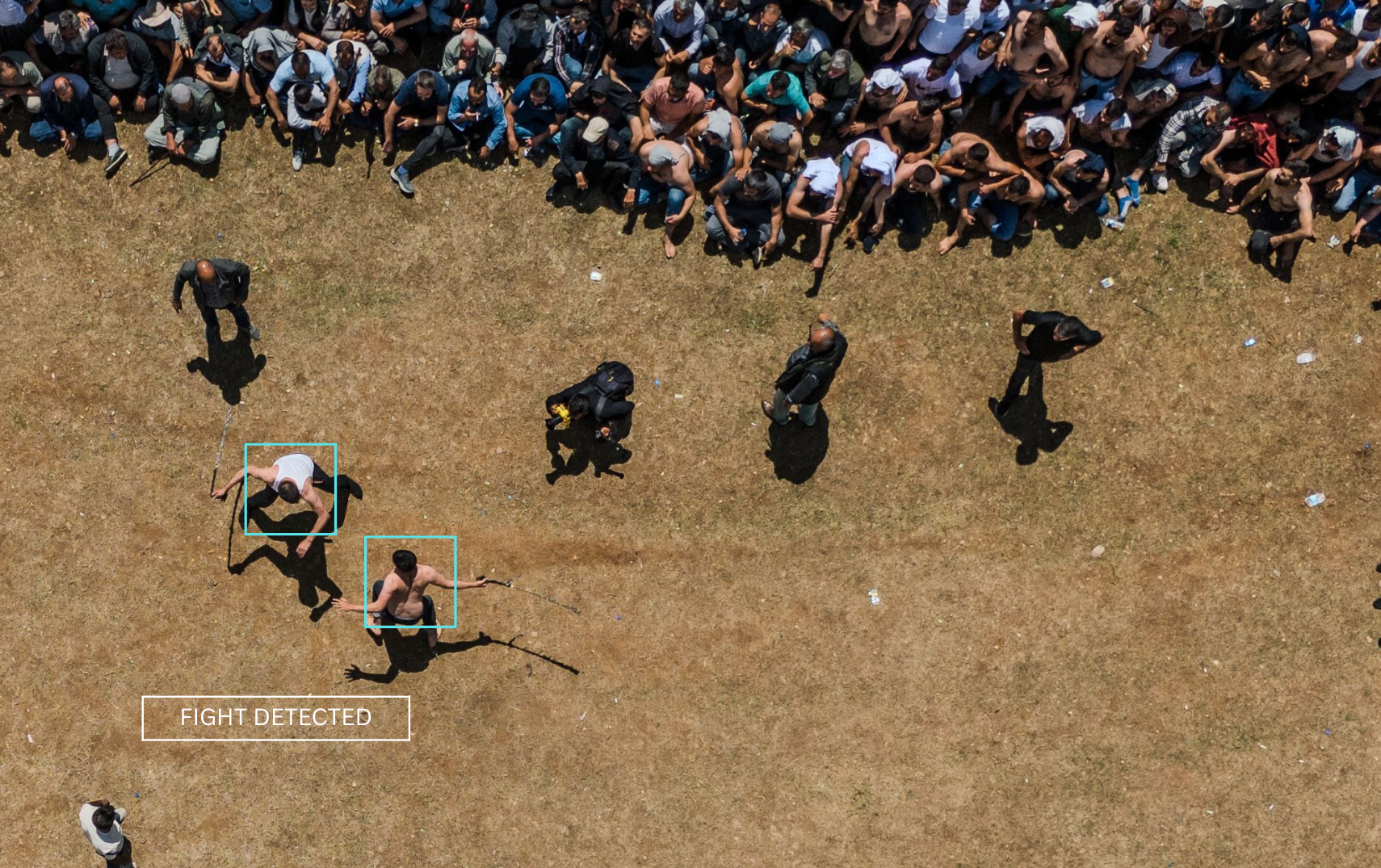 An aerial view of a large crowd of people standing in a line on dry grass. In the center, two fighters are engaged in a physical fight. Several security personnel are nearby, and a box outlines the fight with text overlay 'FIGHT DETECTED.'