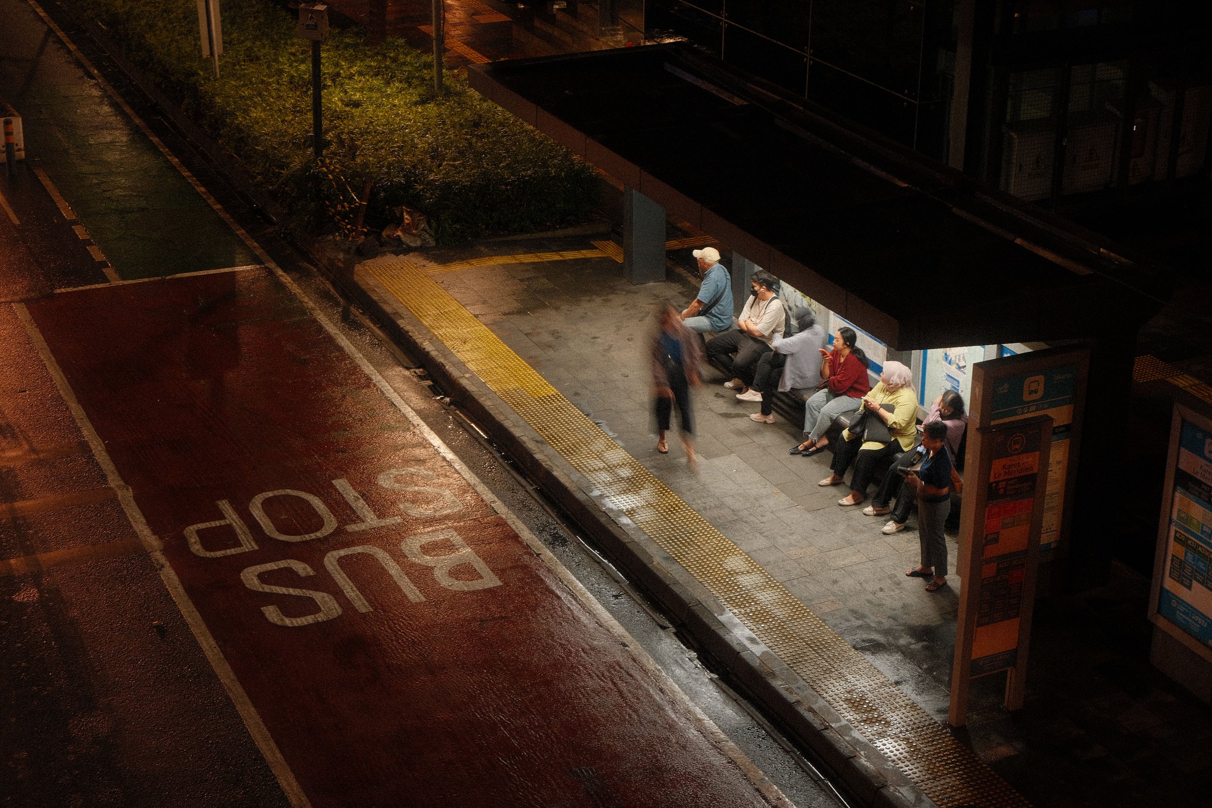 A bus stop shelter at night with several people seated inside. Some are wearing face masks, and one person is looking at their phone. The surroundings are wet, and the street markings are visible on the road.