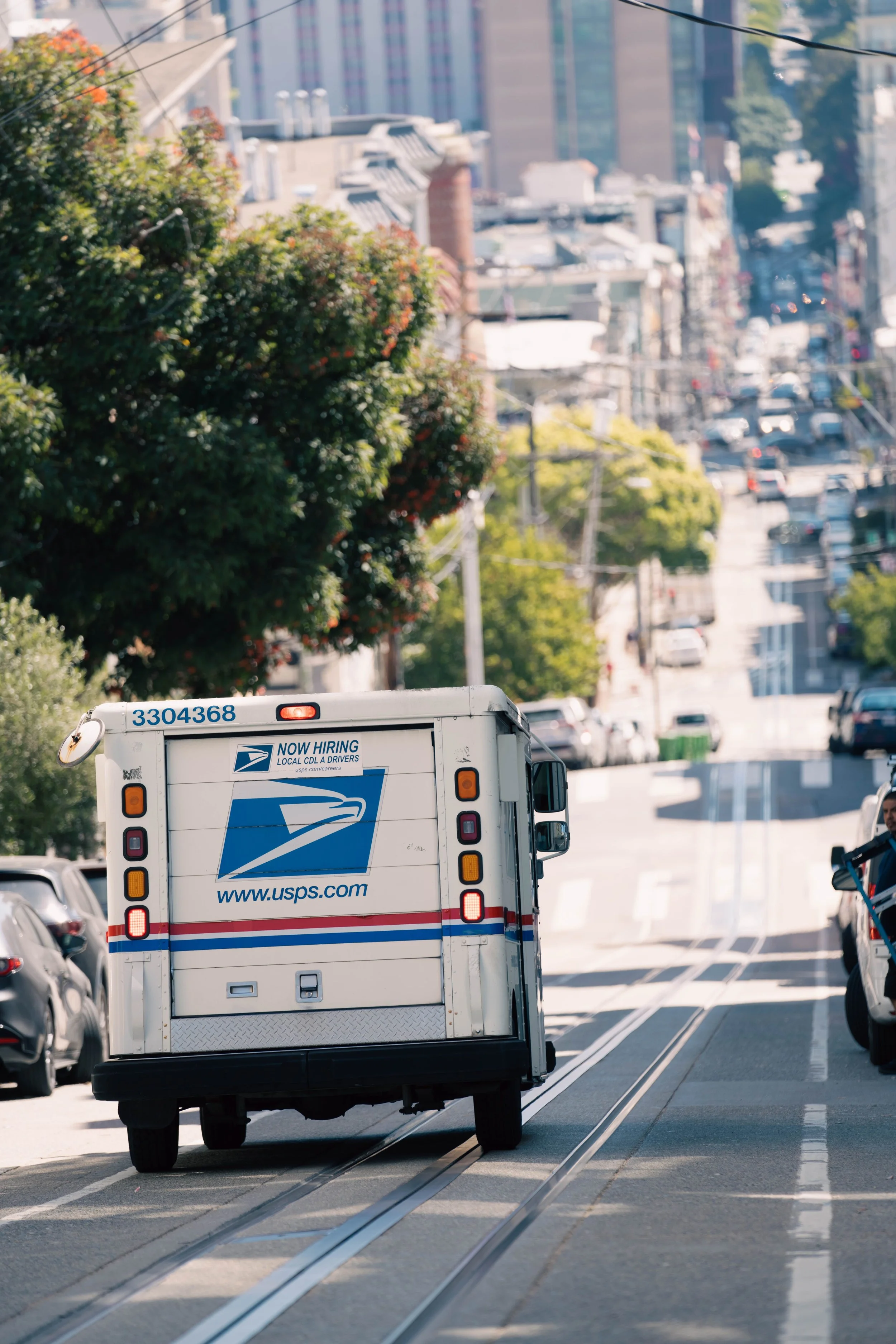 A USPS delivery truck driving down a city street with trolley tracks in the road, lined with parked cars and trees, in a hilly urban area.