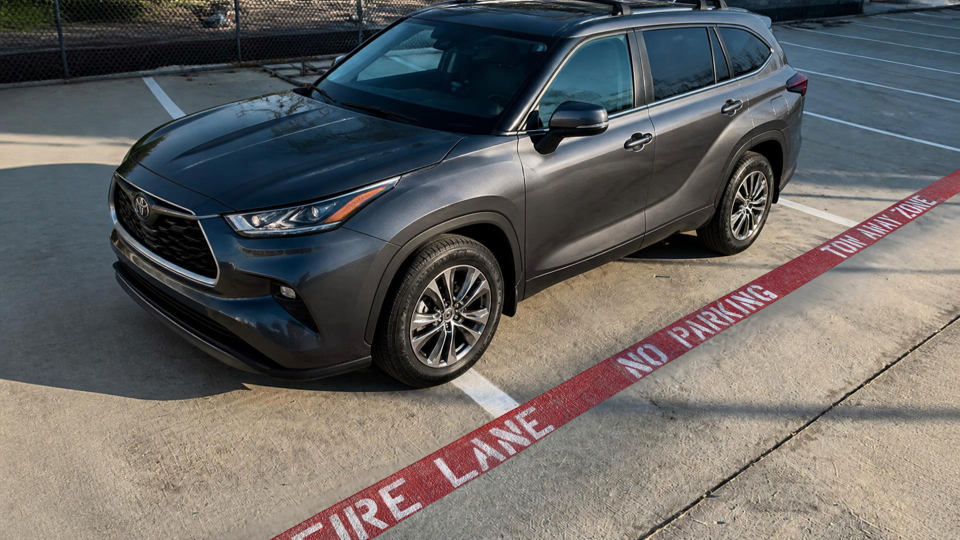 Gray Toyota SUV parked next to a red and white 'Fire Lane No Parking' zone line in an outdoor parking lot with a chain-link fence in the background.