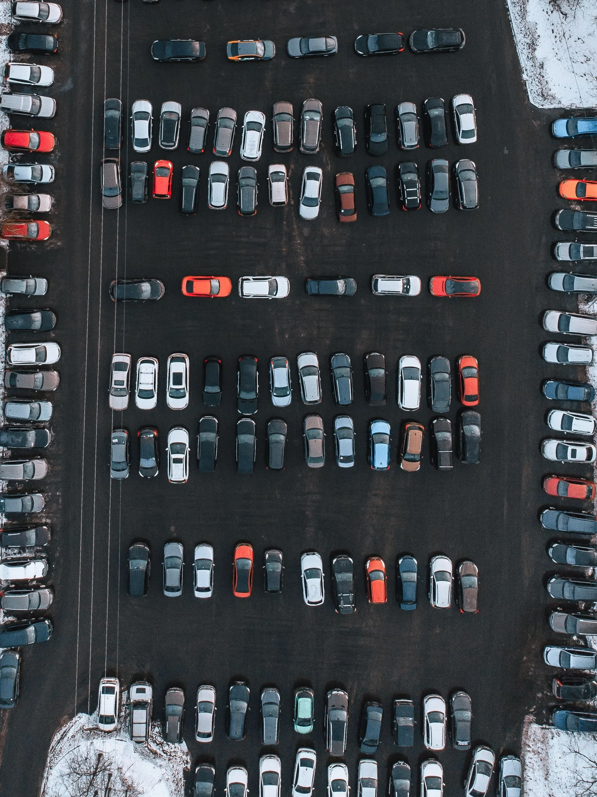 An aerial view of a parking lot with rows of cars parked on asphalt, some snow on the ground.