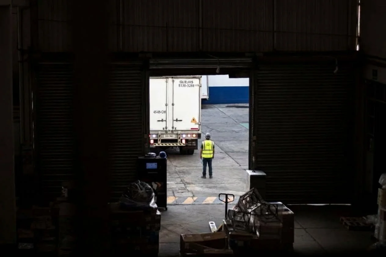 A person wearing a yellow safety vest standing outside a large warehouse door, with a truck parked nearby, seen from inside the warehouse with various boxes and equipment in the foreground.
