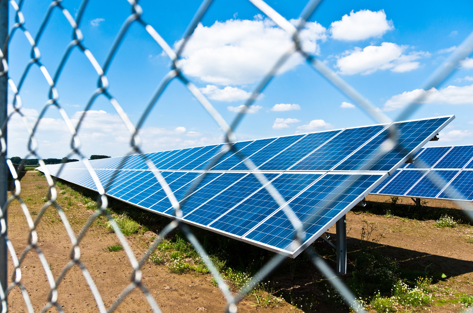 Row of solar panels behind a chain-link fence, under a blue sky with clouds.