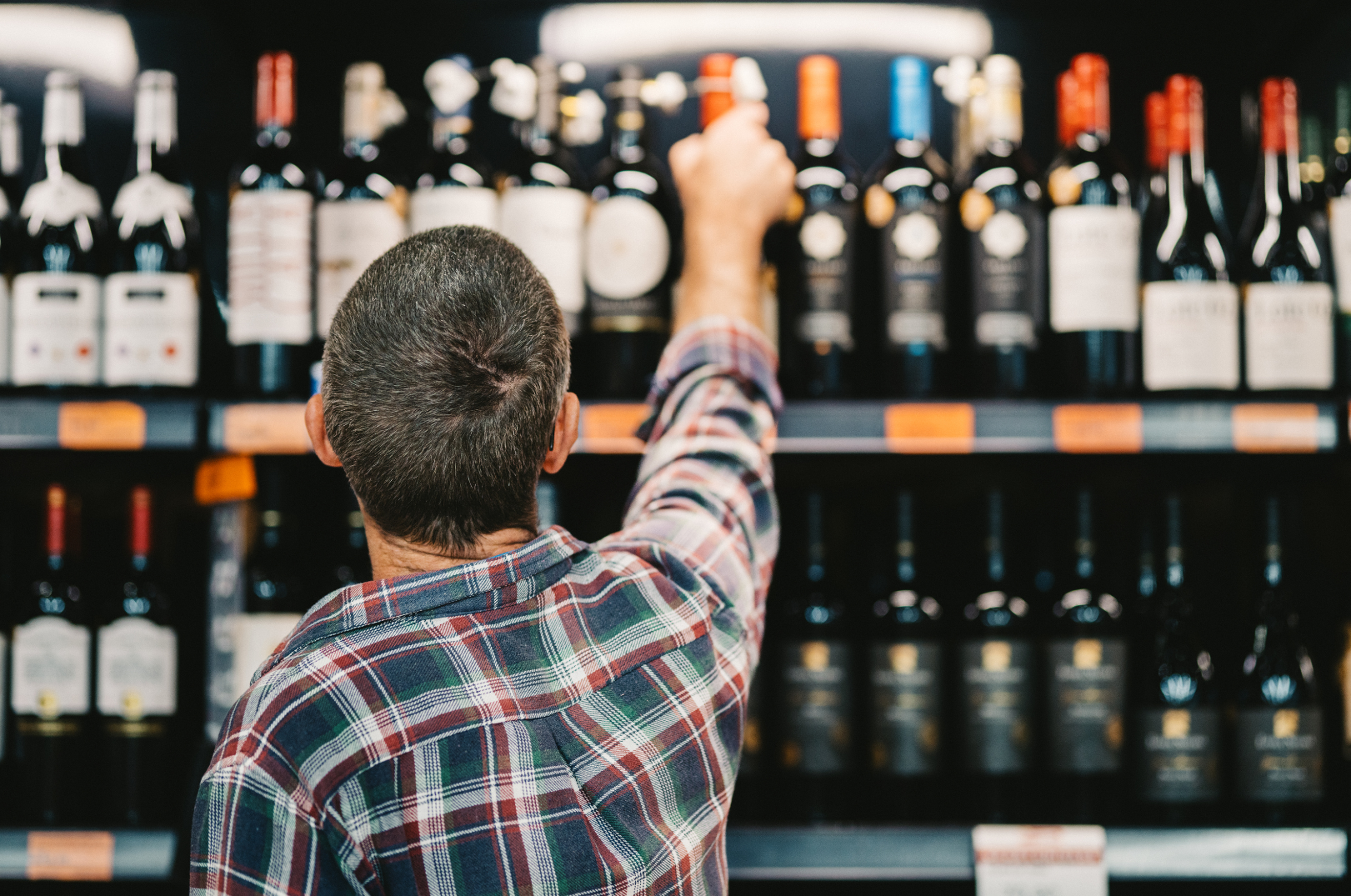 A man in a plaid shirt reaching for a bottle of red wine on store shelf.