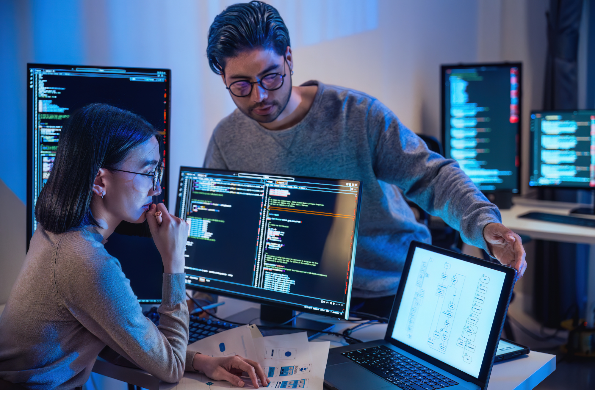 Two individuals, a man and a woman, working on computers in a tech office. The woman is focused on her screen, which displays code, while the man is discussing or explaining something to her. Multiple monitors with code and diagrams are visible in the background.