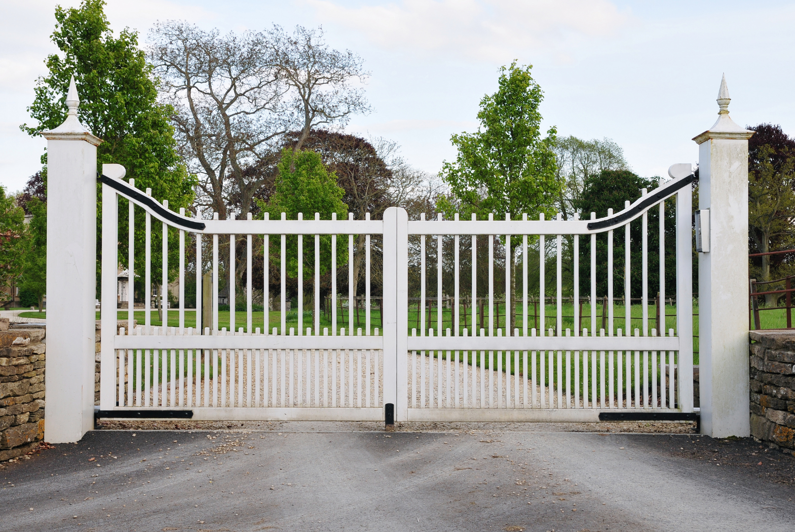 A white metal gate with decorative tops at each side, set between two stone pillars, leading to a green park or garden with trees and a gravel path.