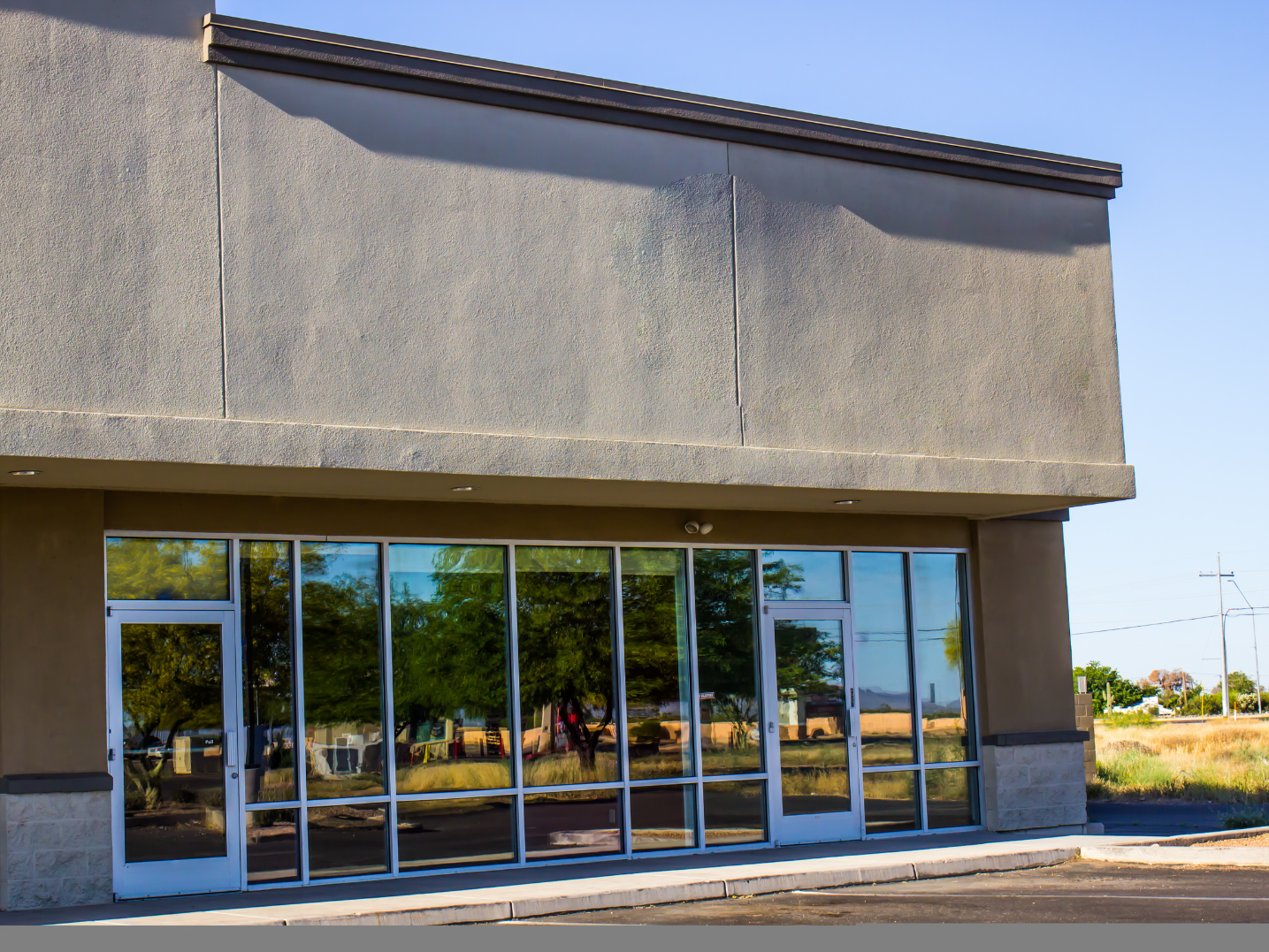 Exterior of an empty commercial building with large glass doors and windows, reflecting trees and the sky, under a clear blue sky.
