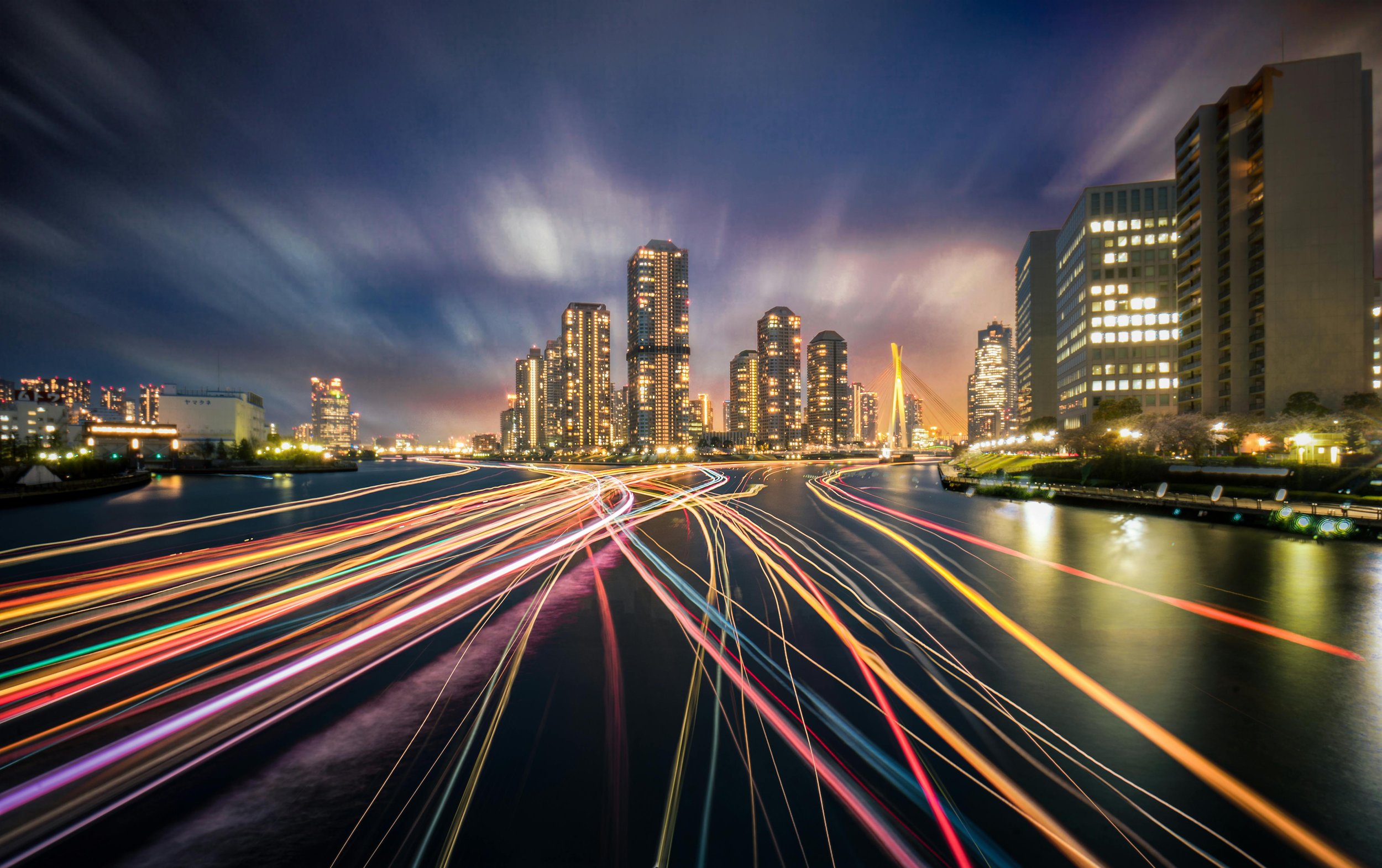 City skyline at night with illuminated skyscrapers, busy roads with light trails from moving vehicles, and a bridge with bright cables, under a partly cloudy sky.